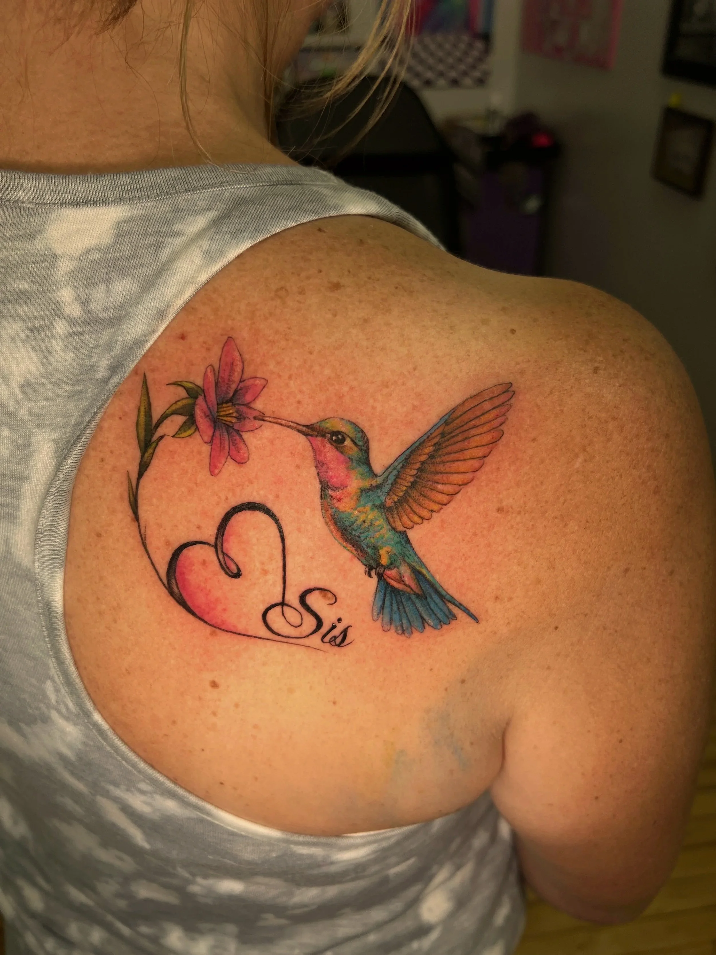 A person's upper arm with a colorful hummingbird tattoo near the shoulder. The tattoo features a hummingbird hovering near a pink flower with green leaves. The person's skin has freckles and they are wearing a grey sleeveless shirt.