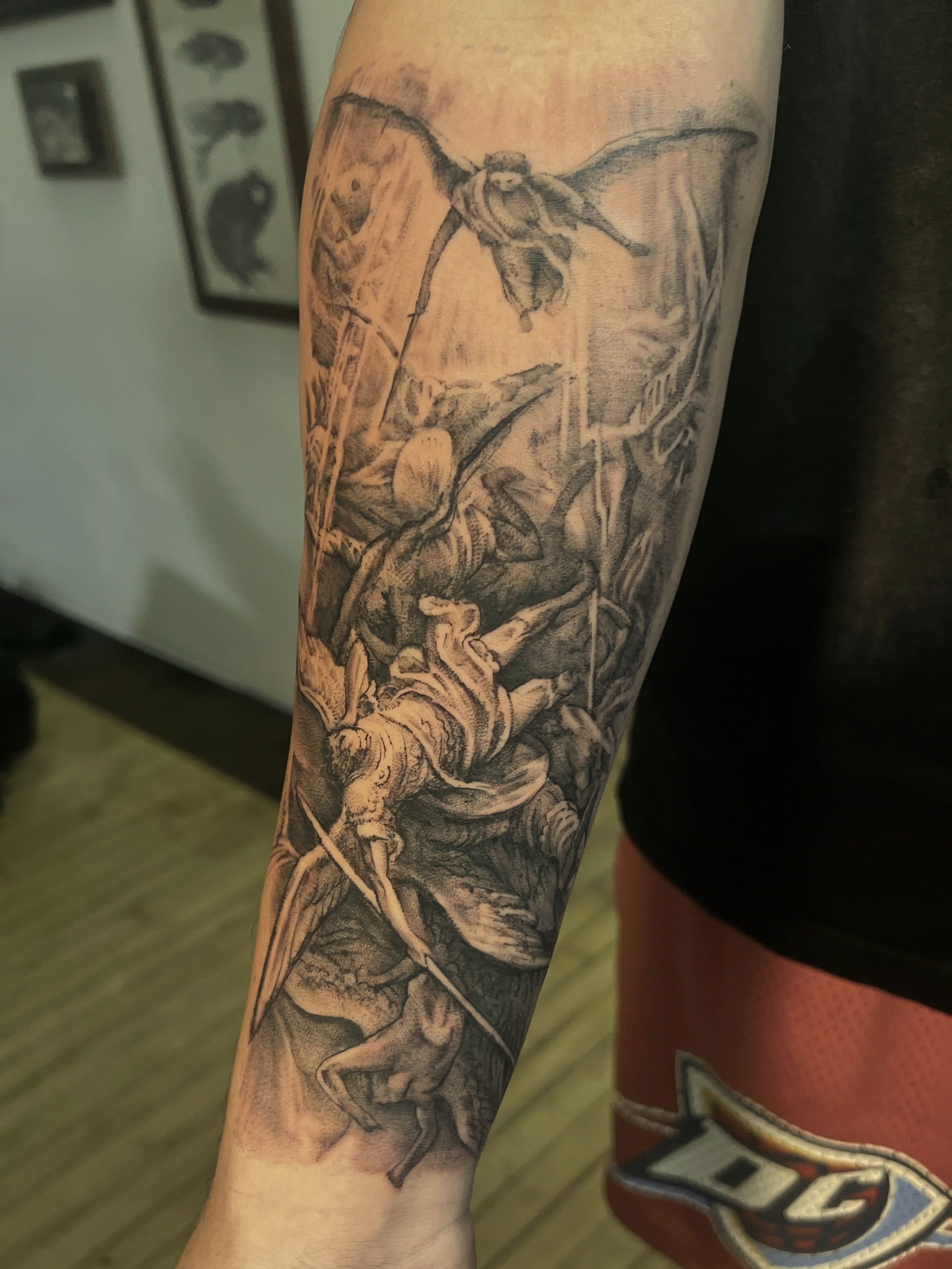 A black and gray tattoo on a person's forearm depicting mythological beings, including a creature with wings and a human figure holding a spear, with detailed shading and intricate design. From Gustav Dore illustration