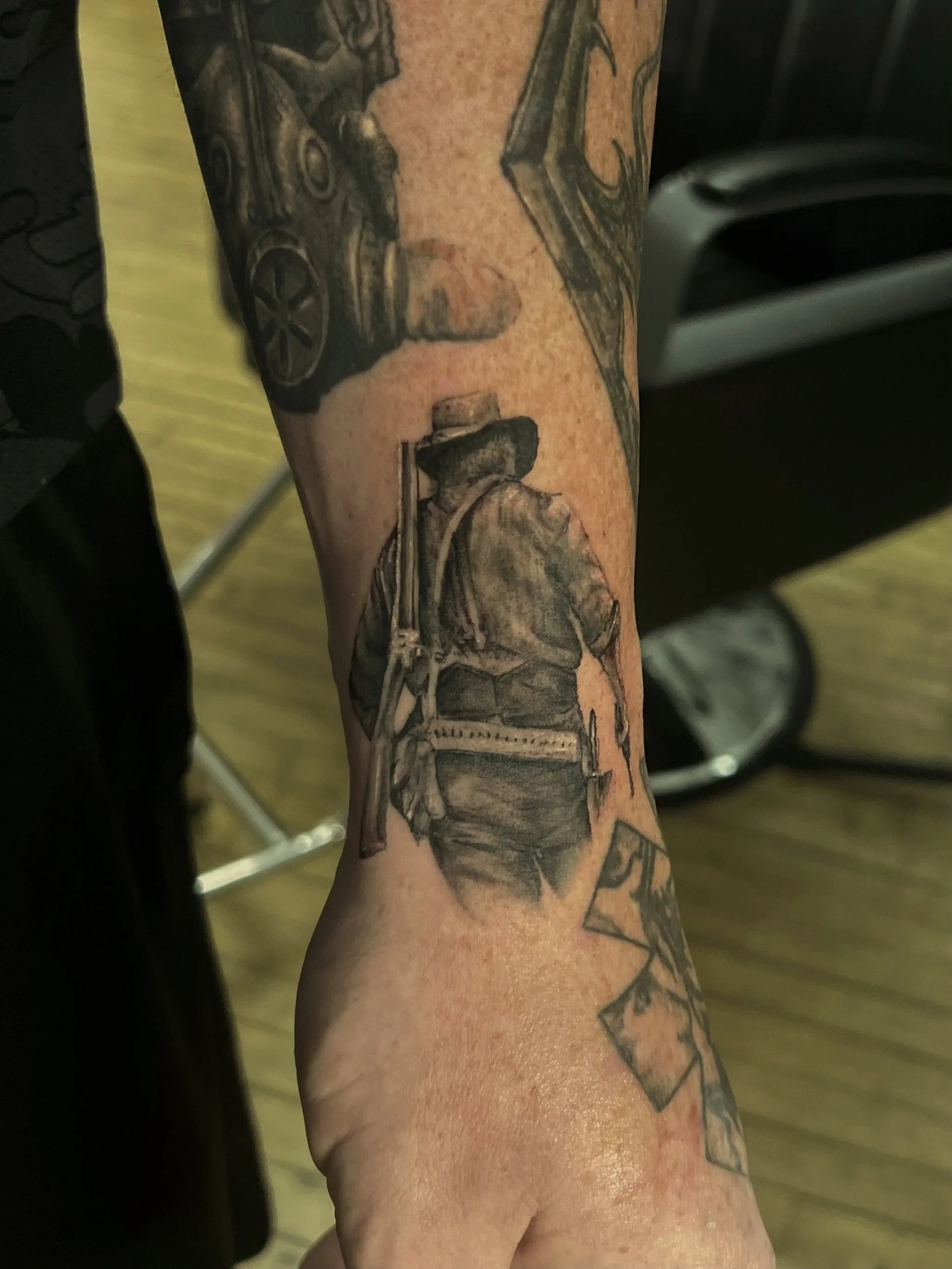 Close-up of a person’s arm with a detailed black and gray tattoo of a person holding a spray can, standing on a chair, with another figure in the background. Reference to Arthur Morgan from Red Dead Redemption