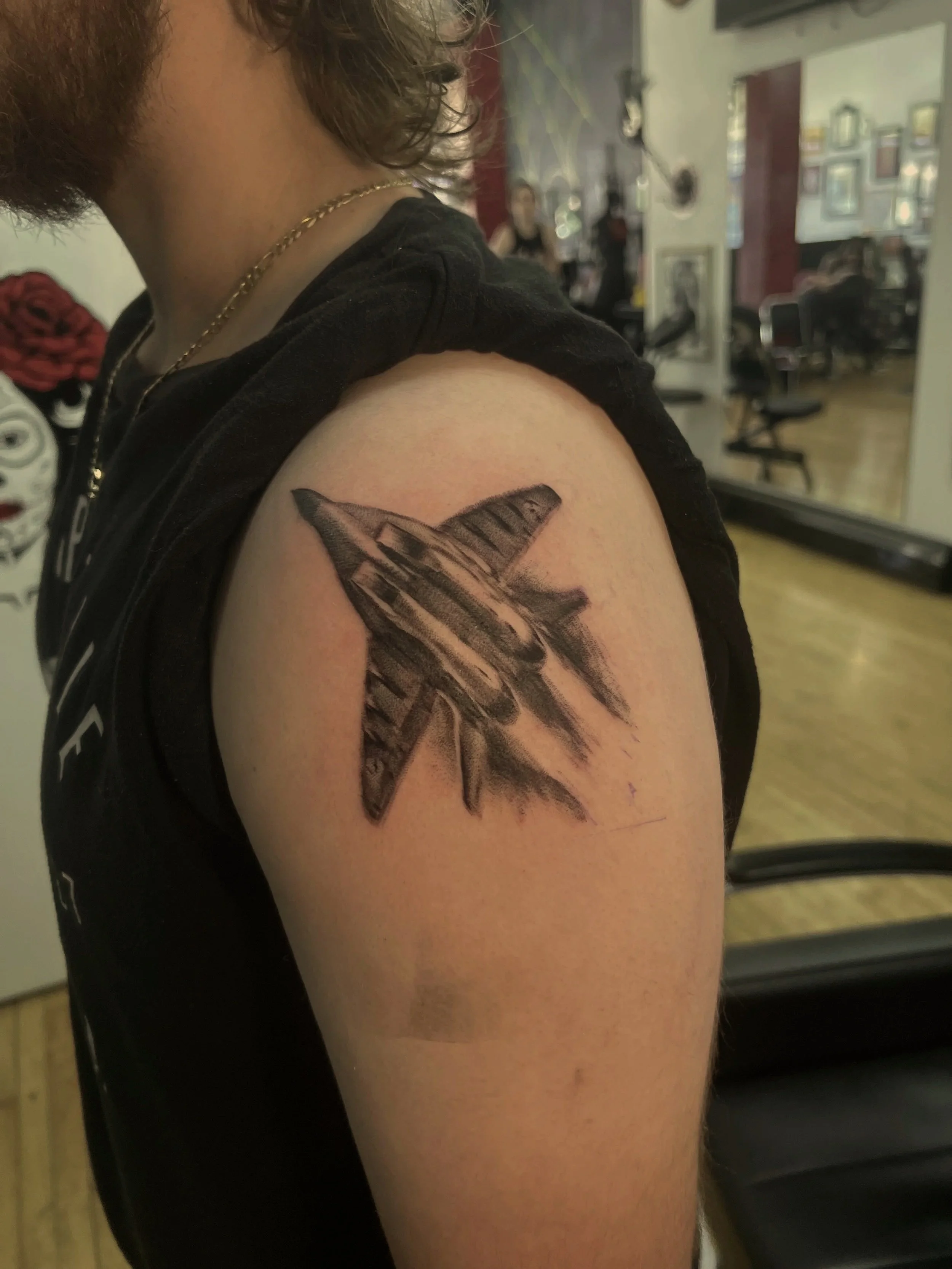 A person with a short curly haircut and a gold chain necklace showing a tattoo of a rocket ship on their upper arm inside a tattoo or art studio.