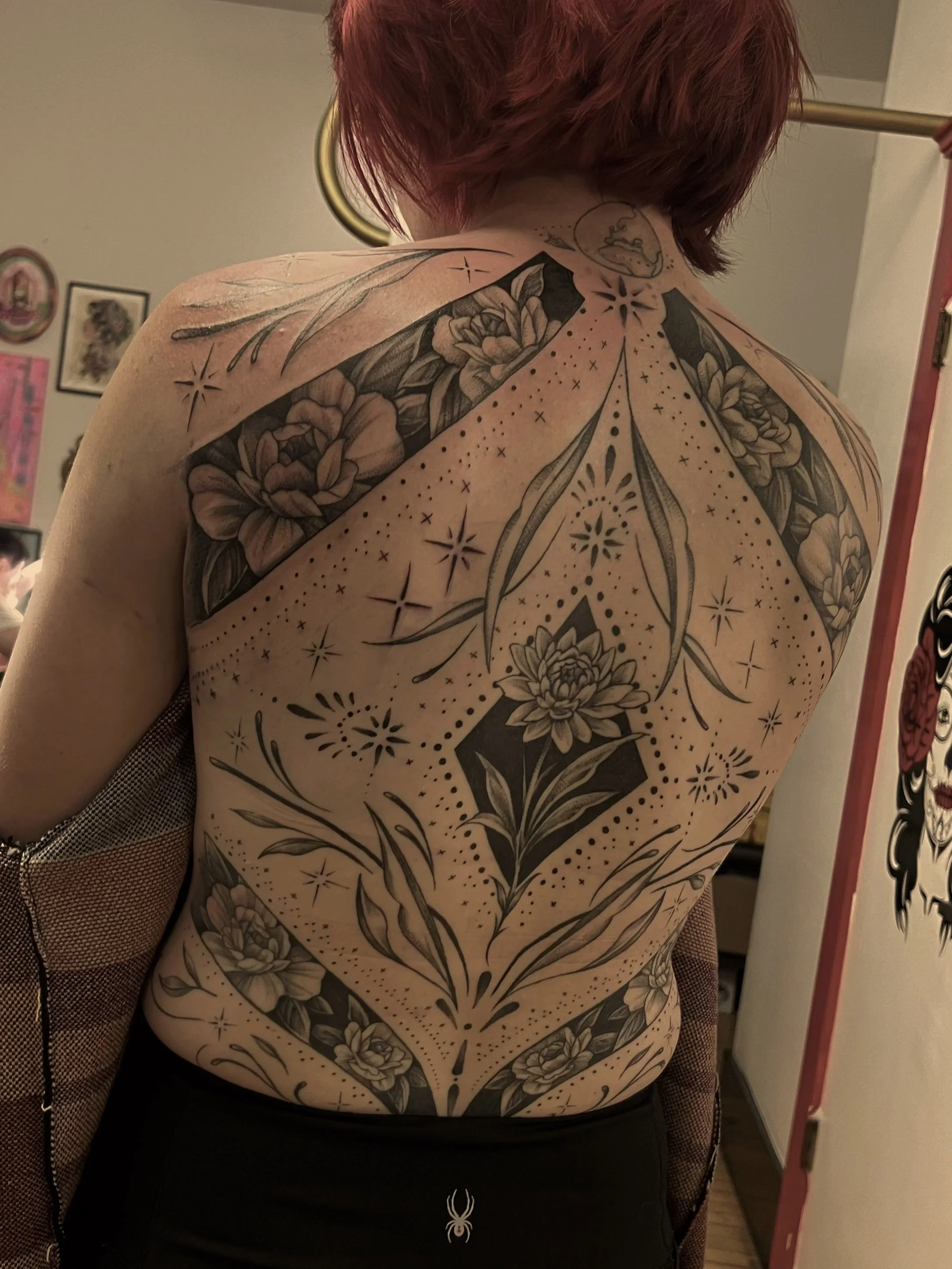 Back view of person with shoulder-length red hair, covered in intricate black and gray floral and geometric tattoos.