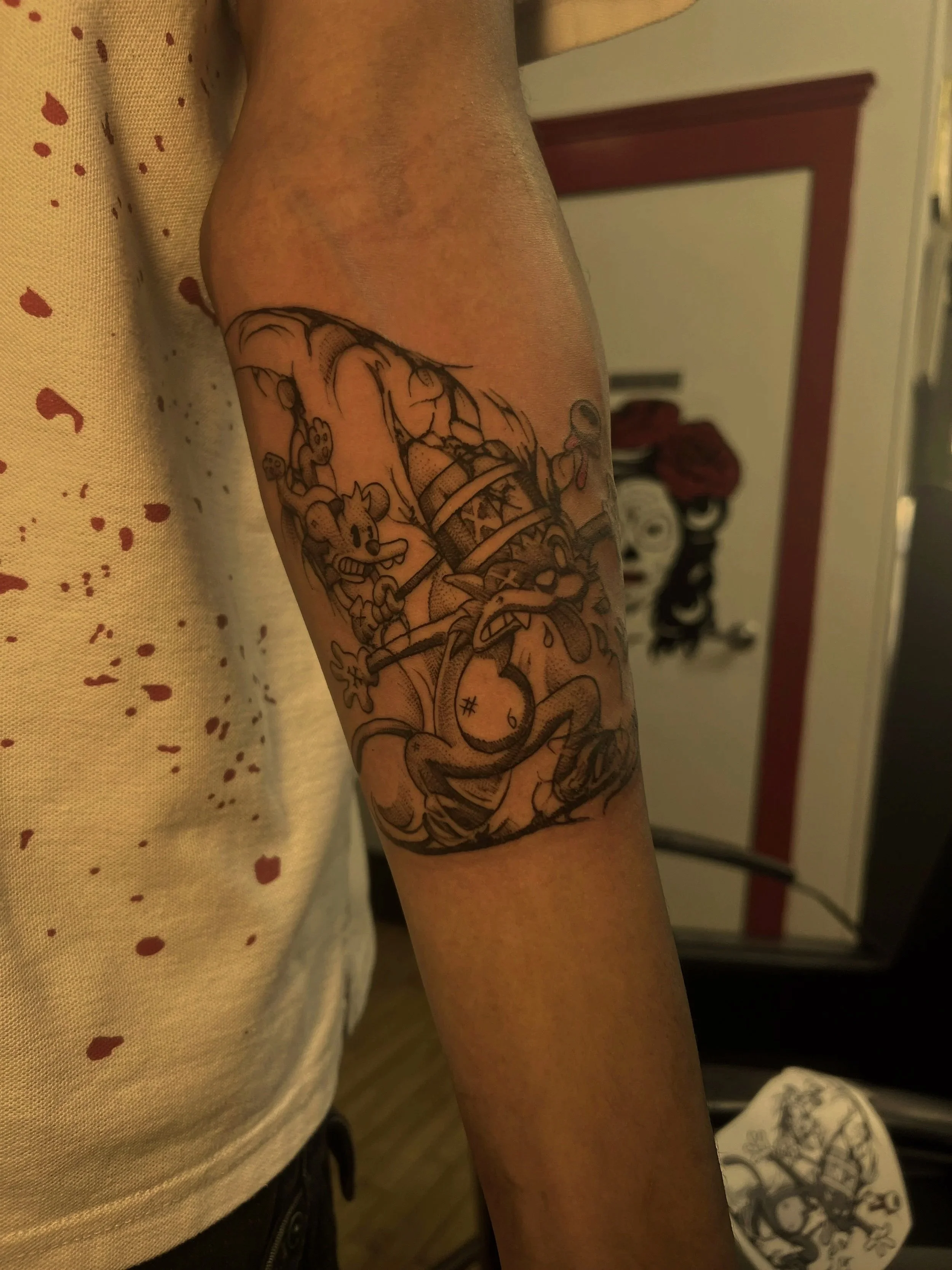 A person's arm with a tattoo of cartoon characters including Mickey Mouse and other Disney characters, with a background of a beige fabric with red splatters and a mirror with a red frame.
