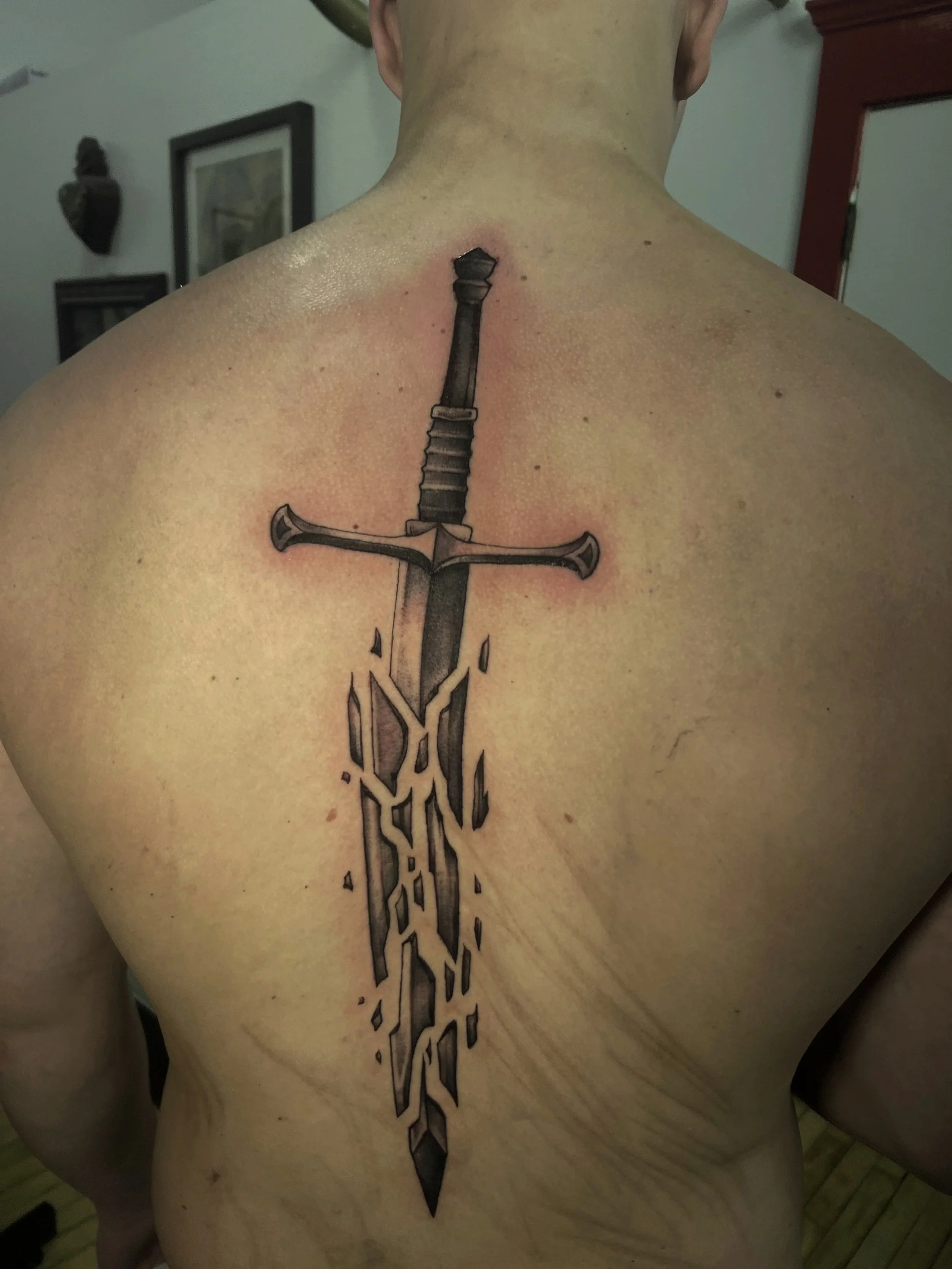 A person's back with a tattoo of a stylized sword running vertically down the spine.