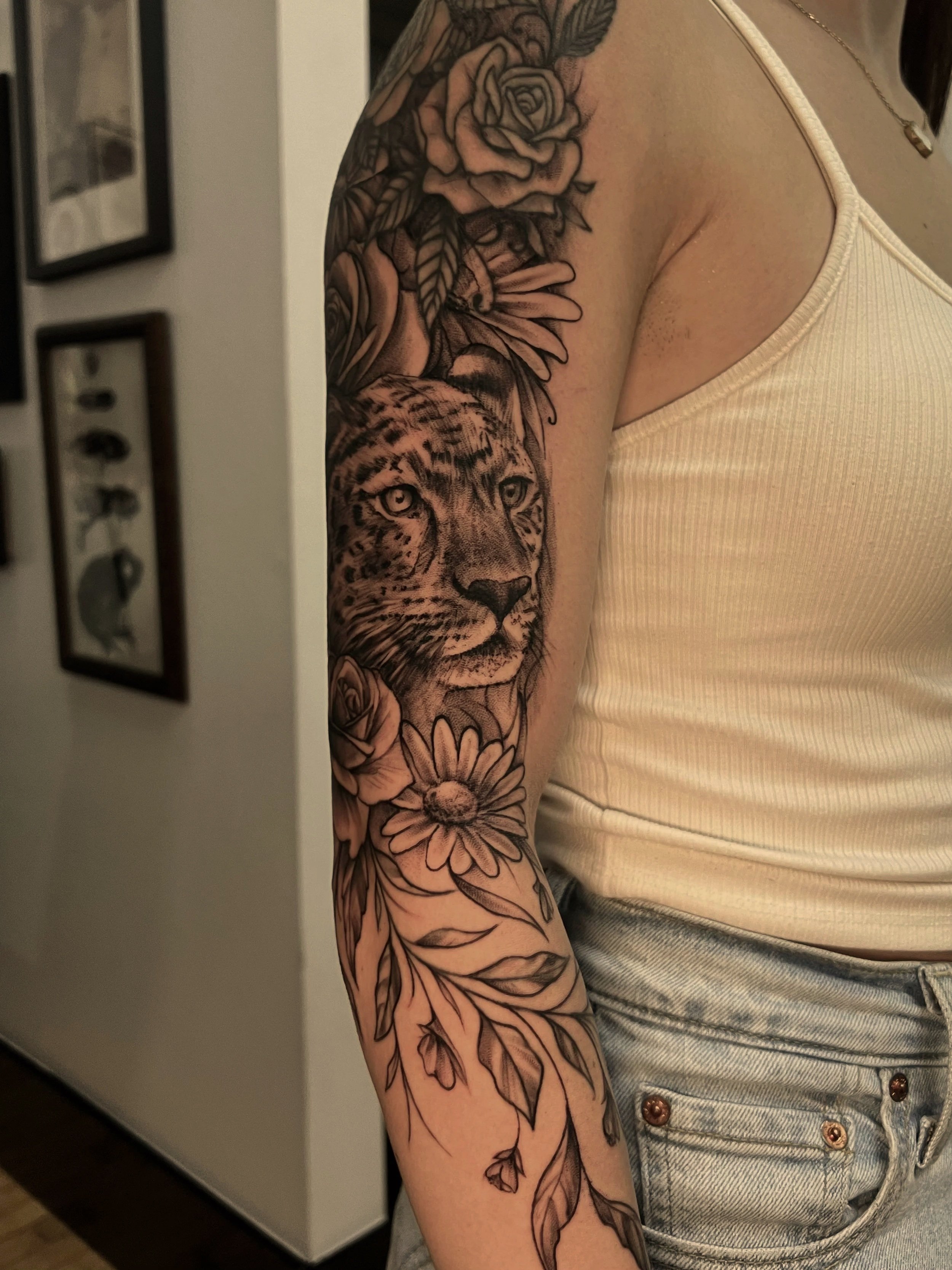 A person's upper arm with a detailed black and gray tattoo of a panther surrounded by flowers and leaves, wearing a cream-colored tank top and light denim shorts.