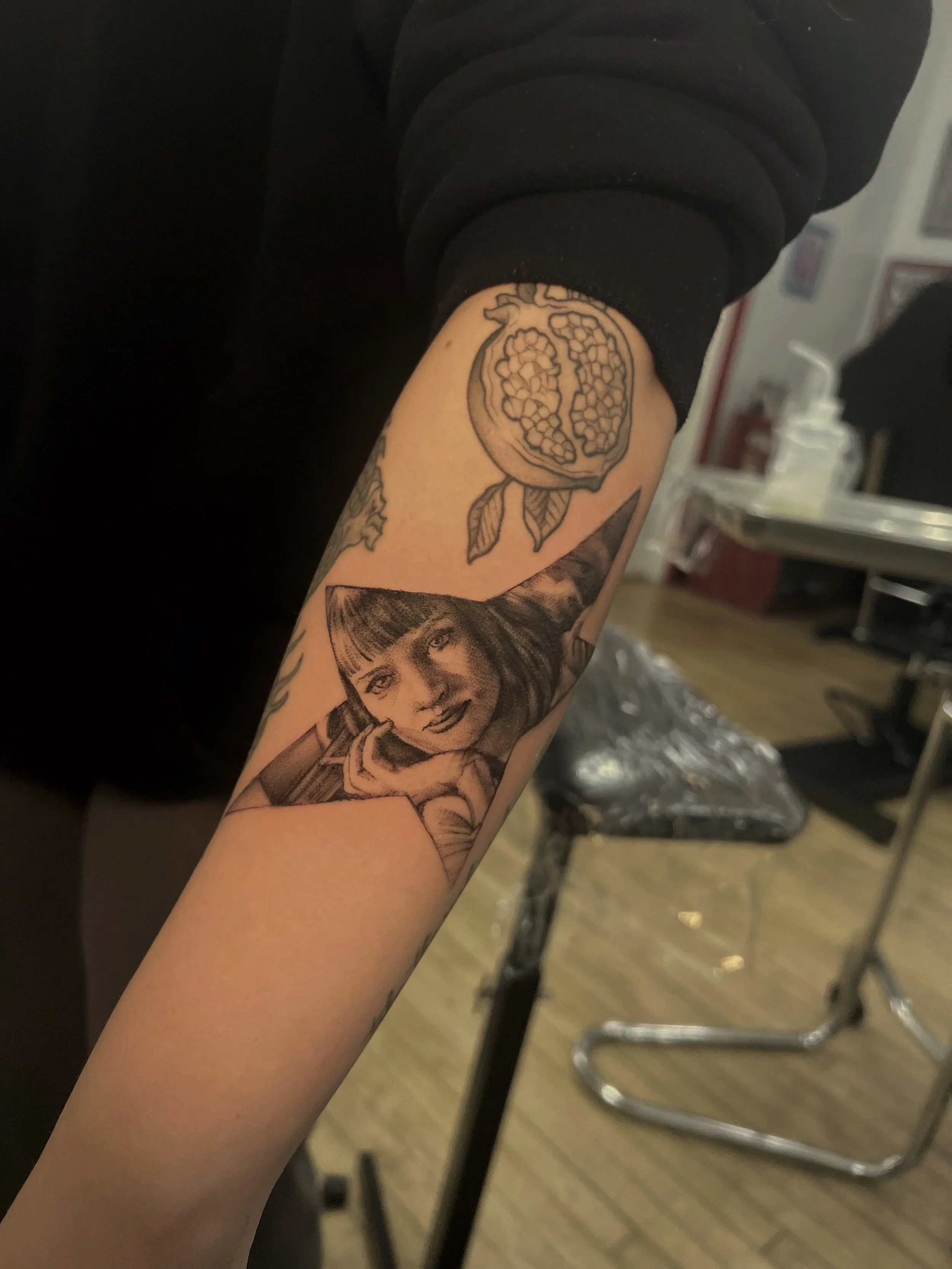 A person with a tattoo of a pomegranate on their forearm, wearing a black sleeve, in a tattoo parlor.