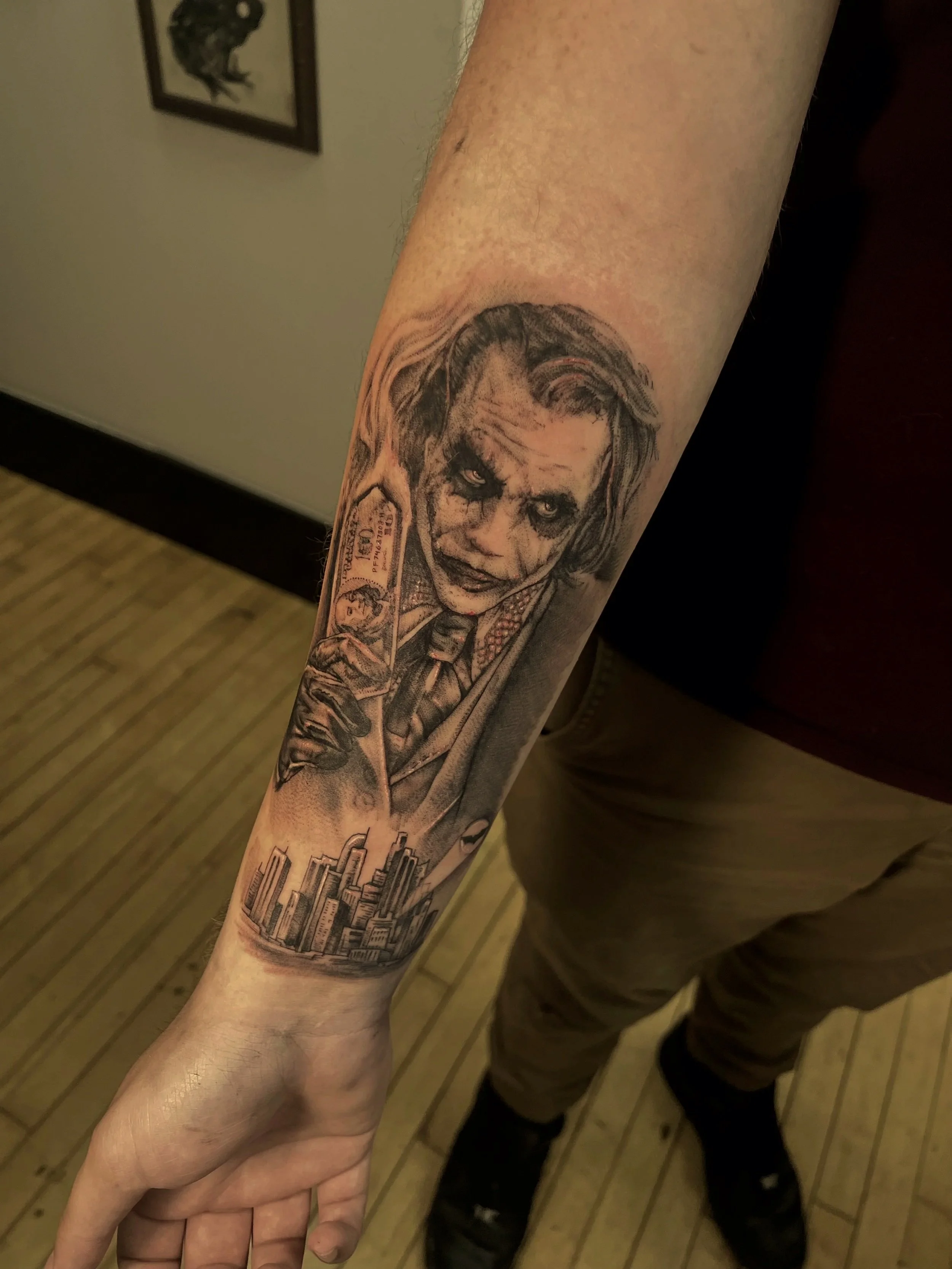 Tattoo of Heath Ledgers Joker from The Dark Knight, with cityscape below on inner forearm.