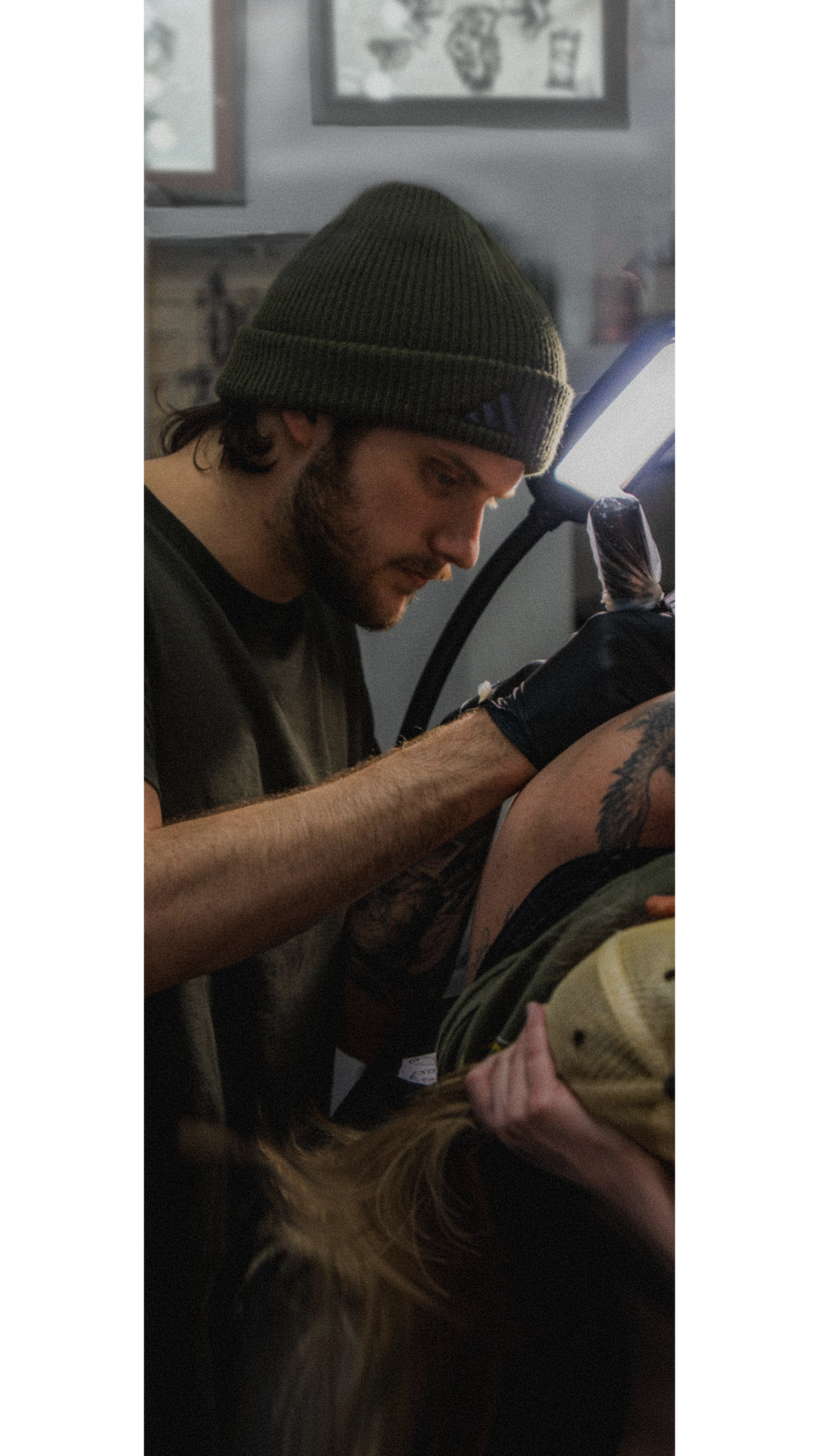 A person with a beard and a green beanie wearing a black shirt is tattooing the arm of another person who is lying down. The background includes framed pictures on the wall and a bright light source.