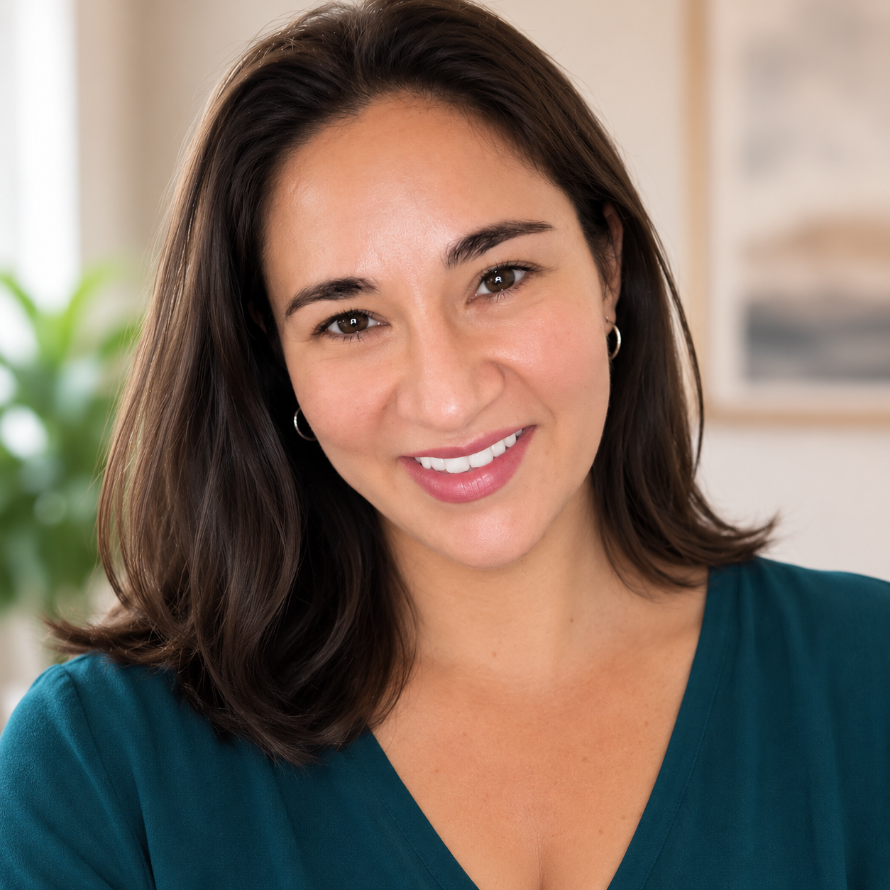 A smiling woman with shoulder-length dark hair wearing a teal top indoors with a plant and framed artwork in the background.