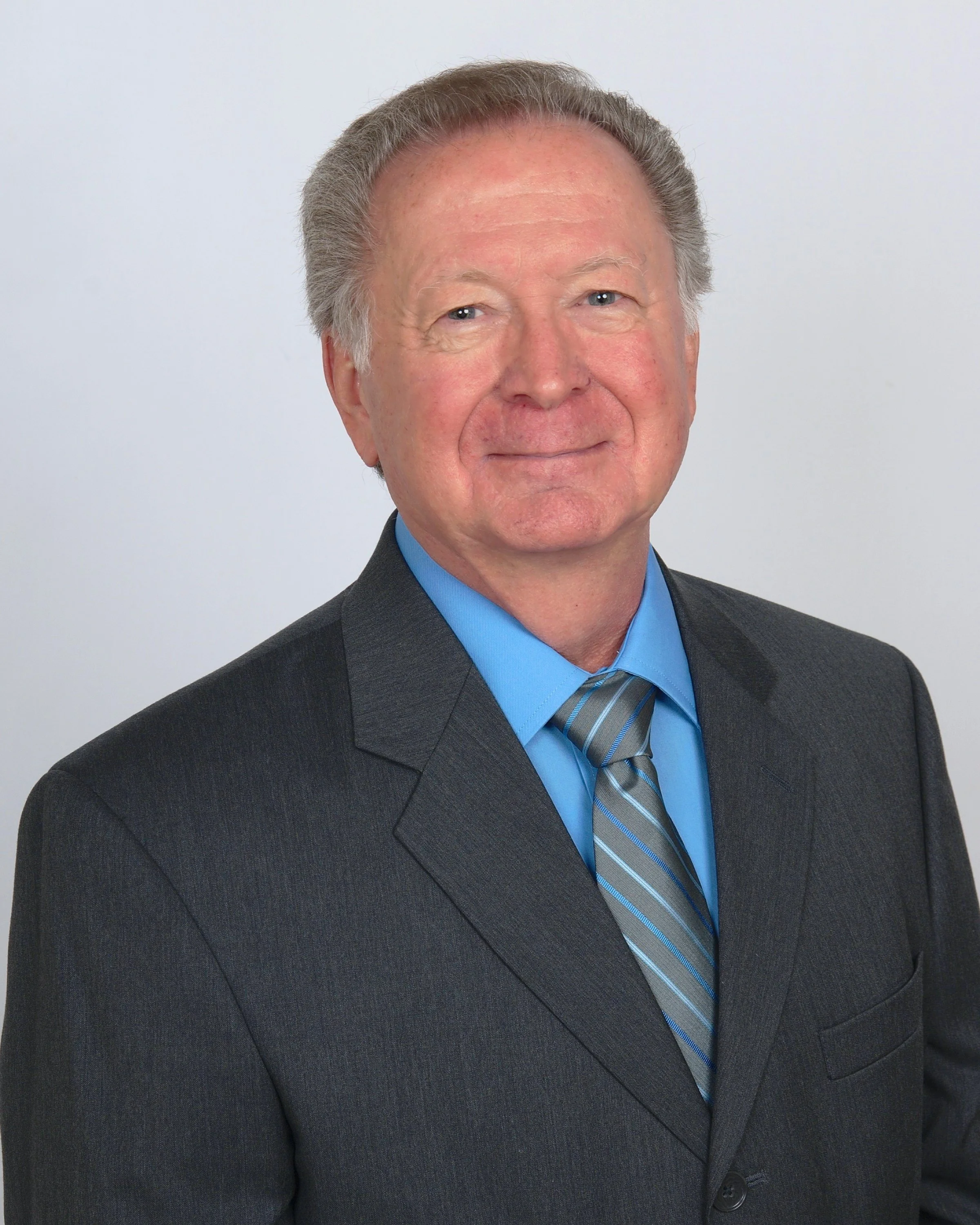 Close-up of a middle-aged man with gray hair, wearing a dark gray suit, blue shirt, and a striped gray and blue tie, standing against a plain light gray background.