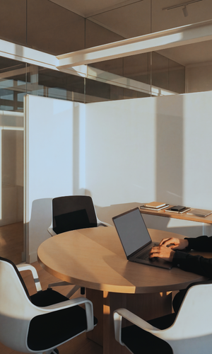 Business meeting room with a round table, laptop, and chairs, with glass walls and modern decor.