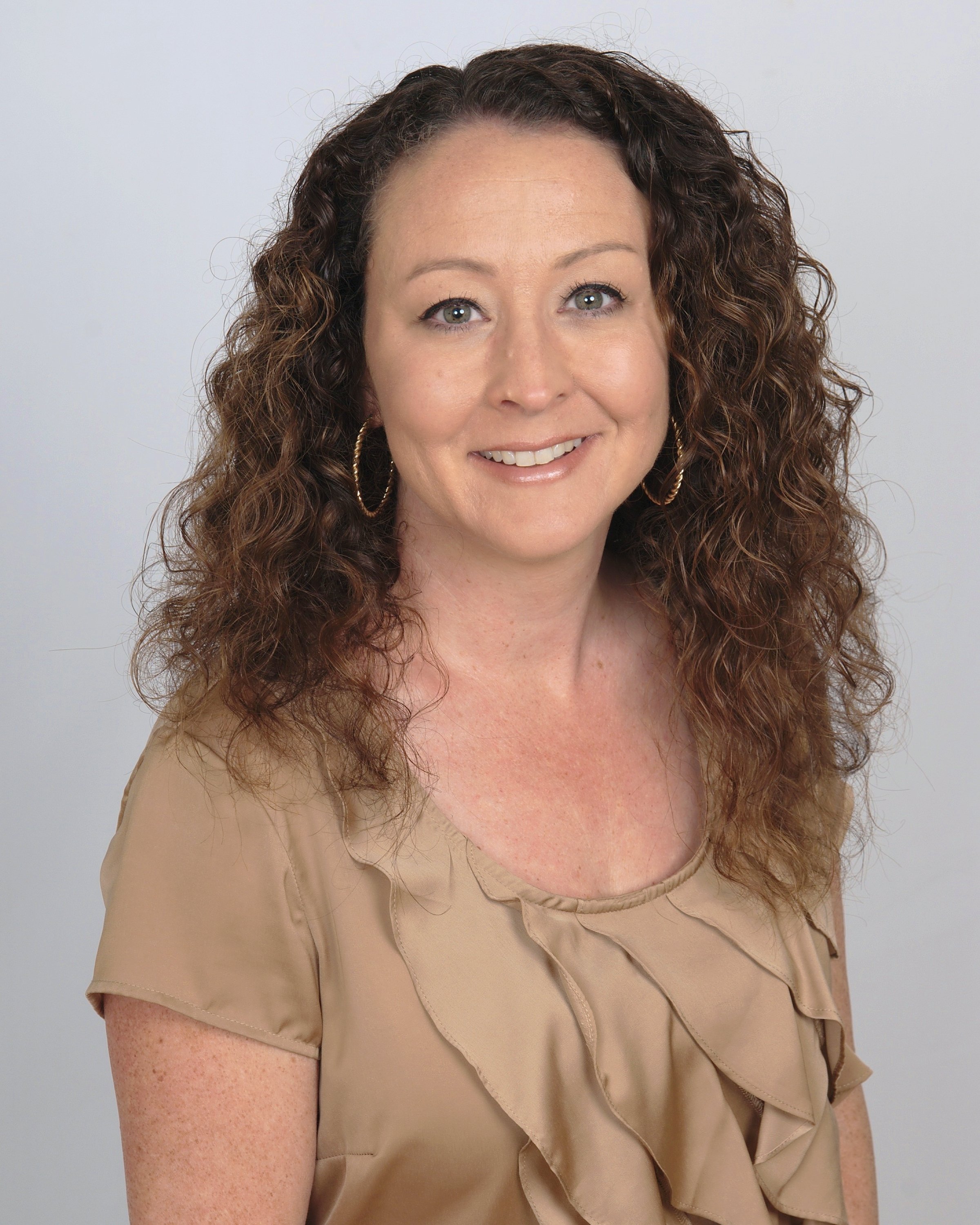 A woman with curly brown hair and hoop earrings smiling at the camera, wearing a beige top with ruffles, against a plain white background.