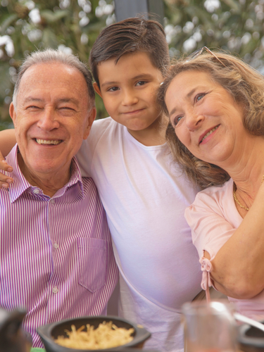 A smiling elderly man and woman with a young boy in between them, outdoors with trees in the background.
