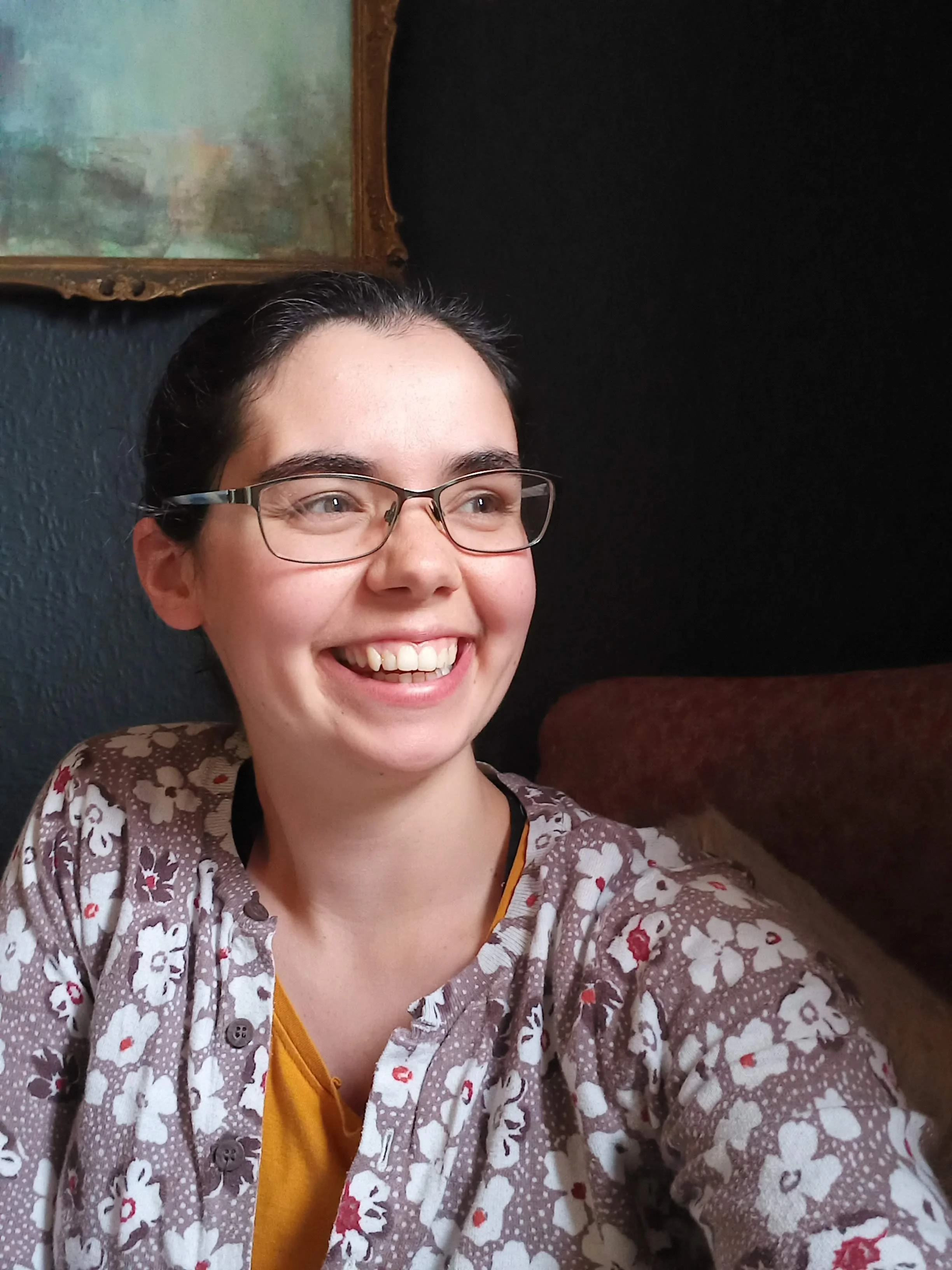 A woman with short dark hair and glasses smiling, taking a selfie in a room with dark walls and a vintage painting hanging behind her.
