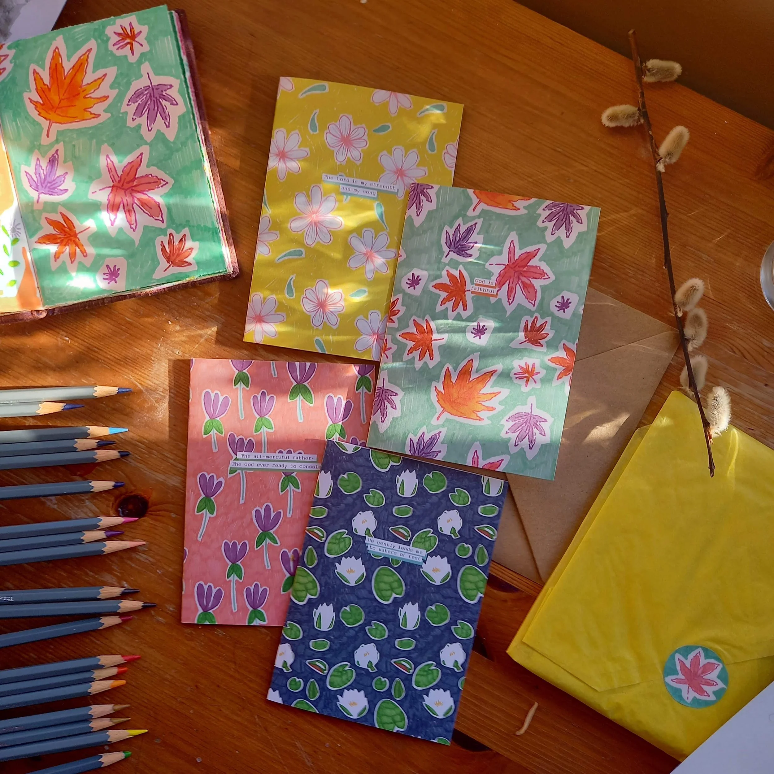 Colorful paper cards with floral and leaf patterns are spread on a wooden table beside colored pencils and a yellow packaging.