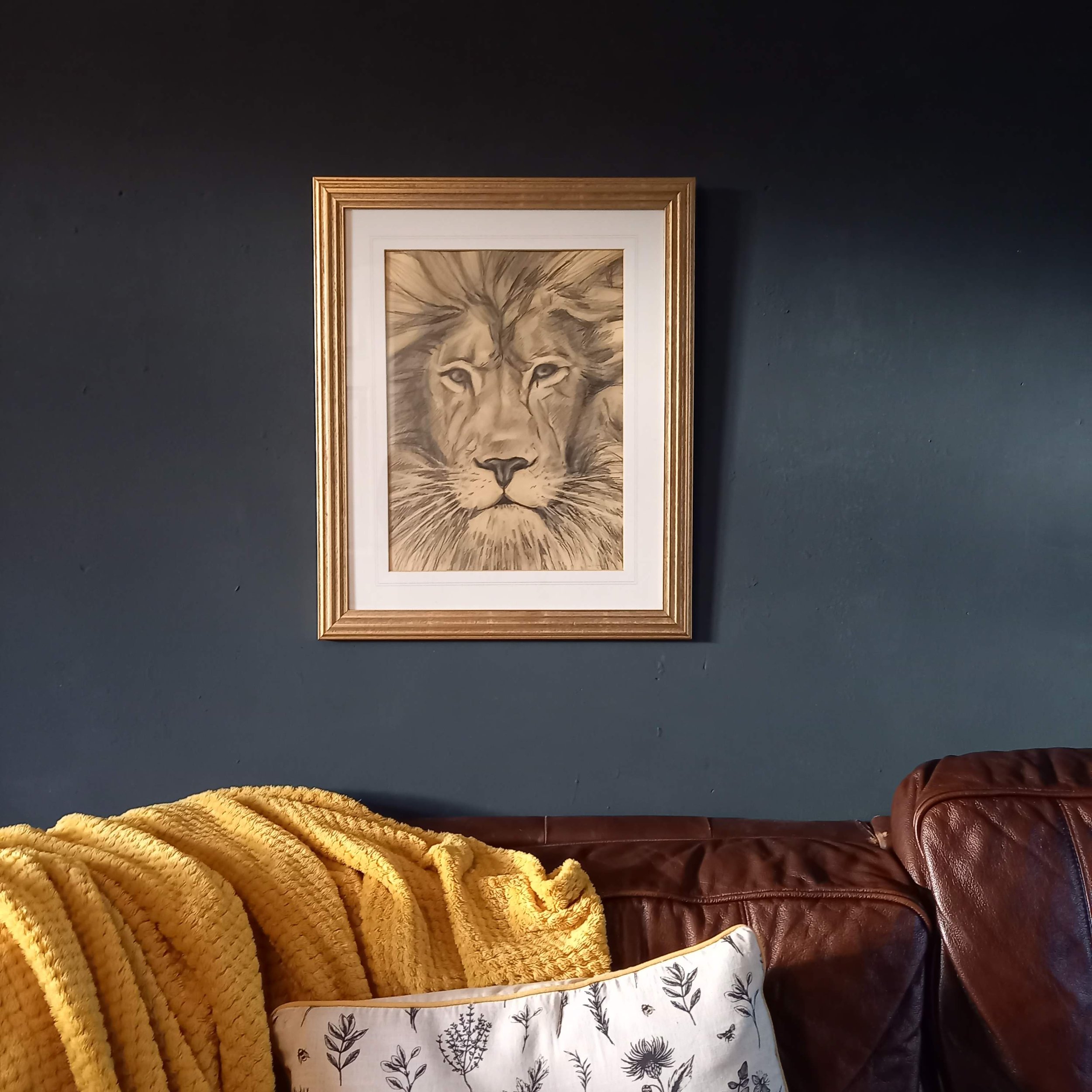 Part of a framed drawing of a lion's face, on a dark wall with a mustard yellow blanket and a leather bag and a floral-patterned fabric visible in the foreground.