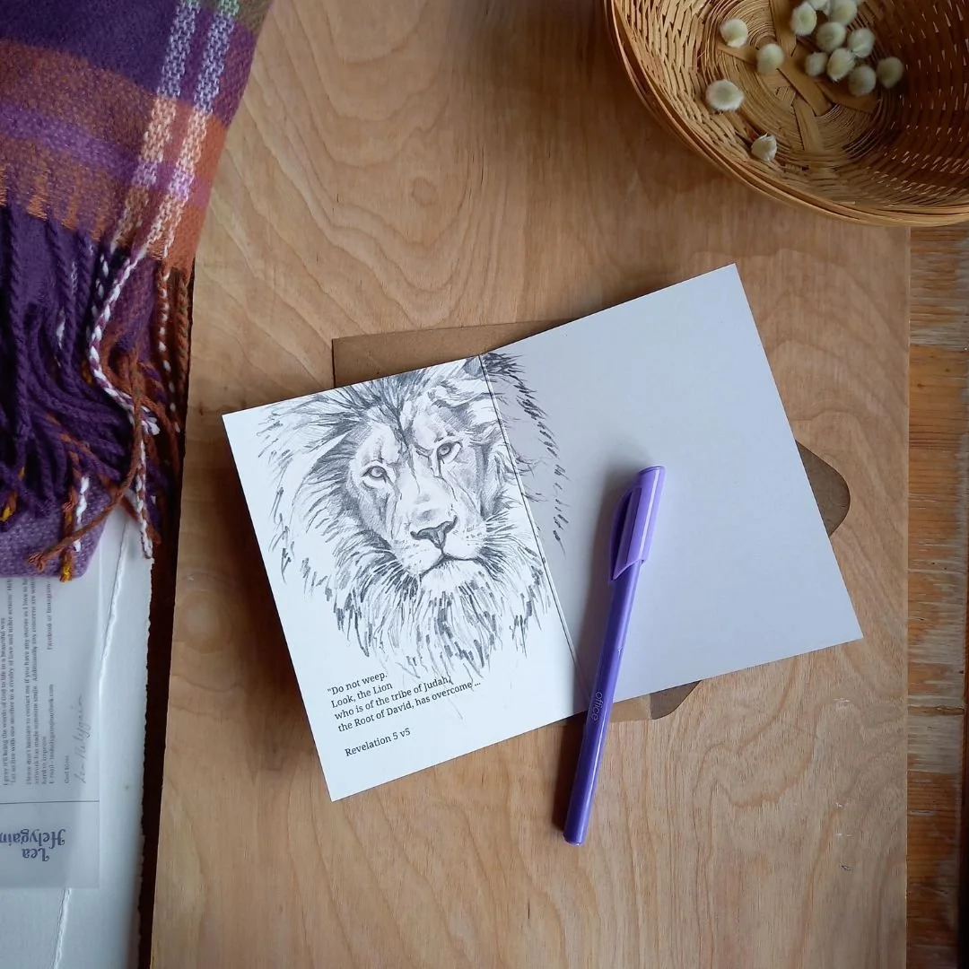 A sketch of a lion's face on a piece of paper with a purple pen, placed on a wooden table next to a basket of small round objects and a folded striped cloth.