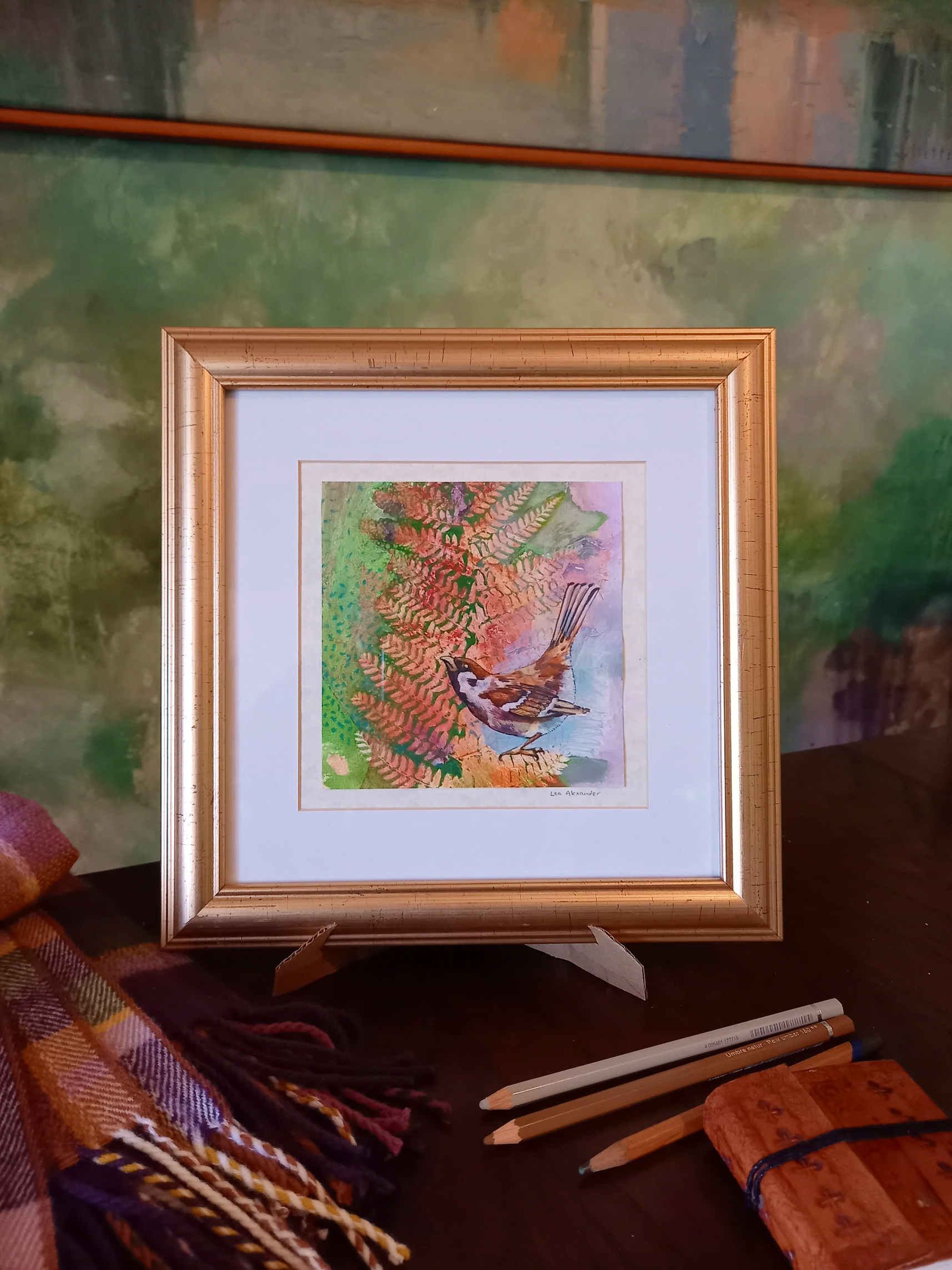 A framed watercolor painting of a bird perched on a branch surrounded by colorful foliage, on a table with colored pencils, a scarf, and a leather notebook.