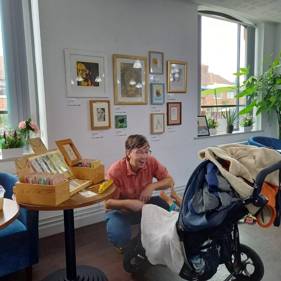 A man sitting on the floor smiling and engaging with a baby in a stroller inside a brightly lit room with a wall decorated with various framed artworks and large windows with plants outside.