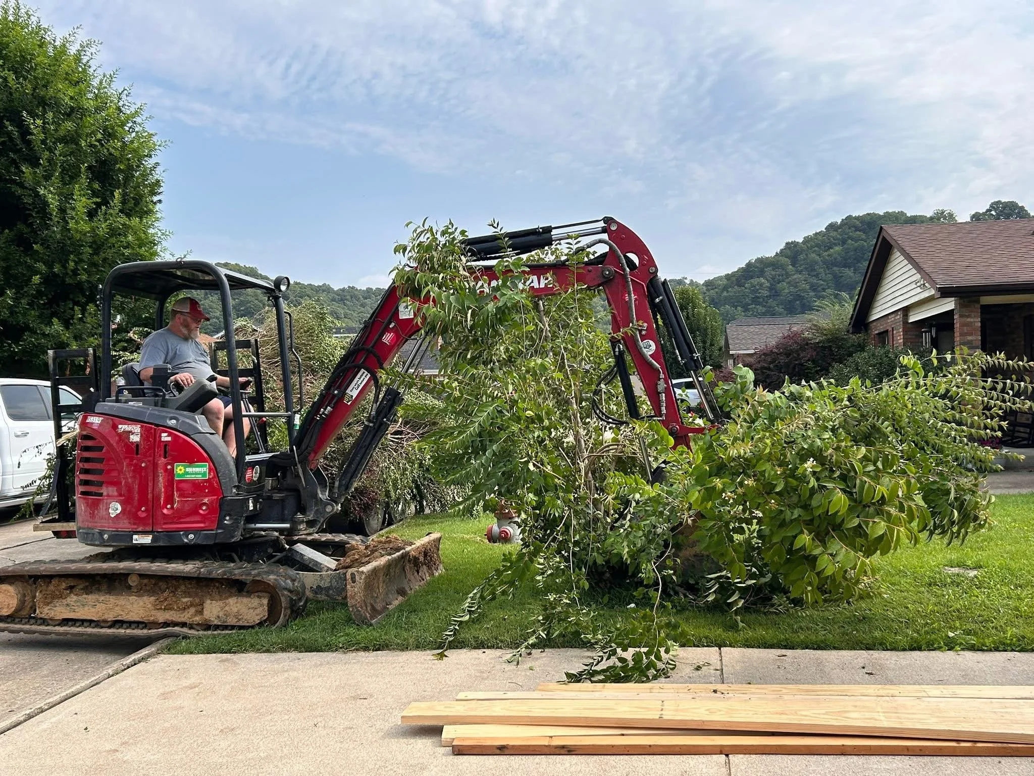 A man operating a small red excavator machine to trim or remove a tree branch in a suburban neighborhood yard, with houses and green hills in the background, and a concrete driveway with wooden planks in the foreground.