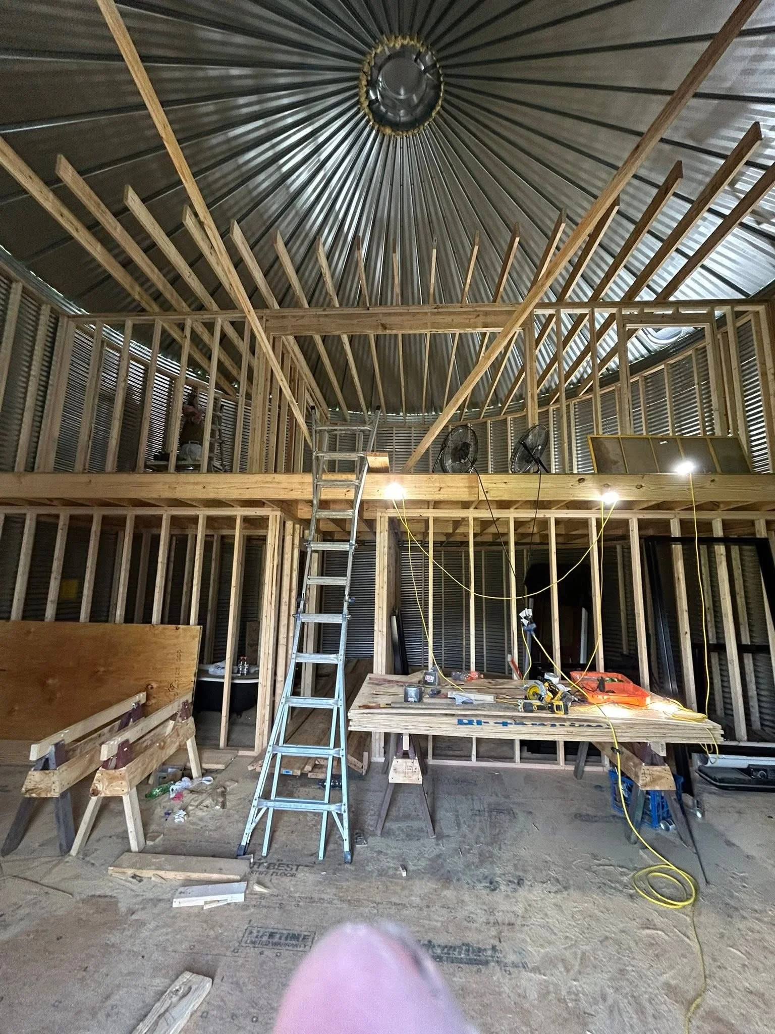 Interior of a building under construction with exposed wooden framing, metal roof, construction tools, and a ladder.