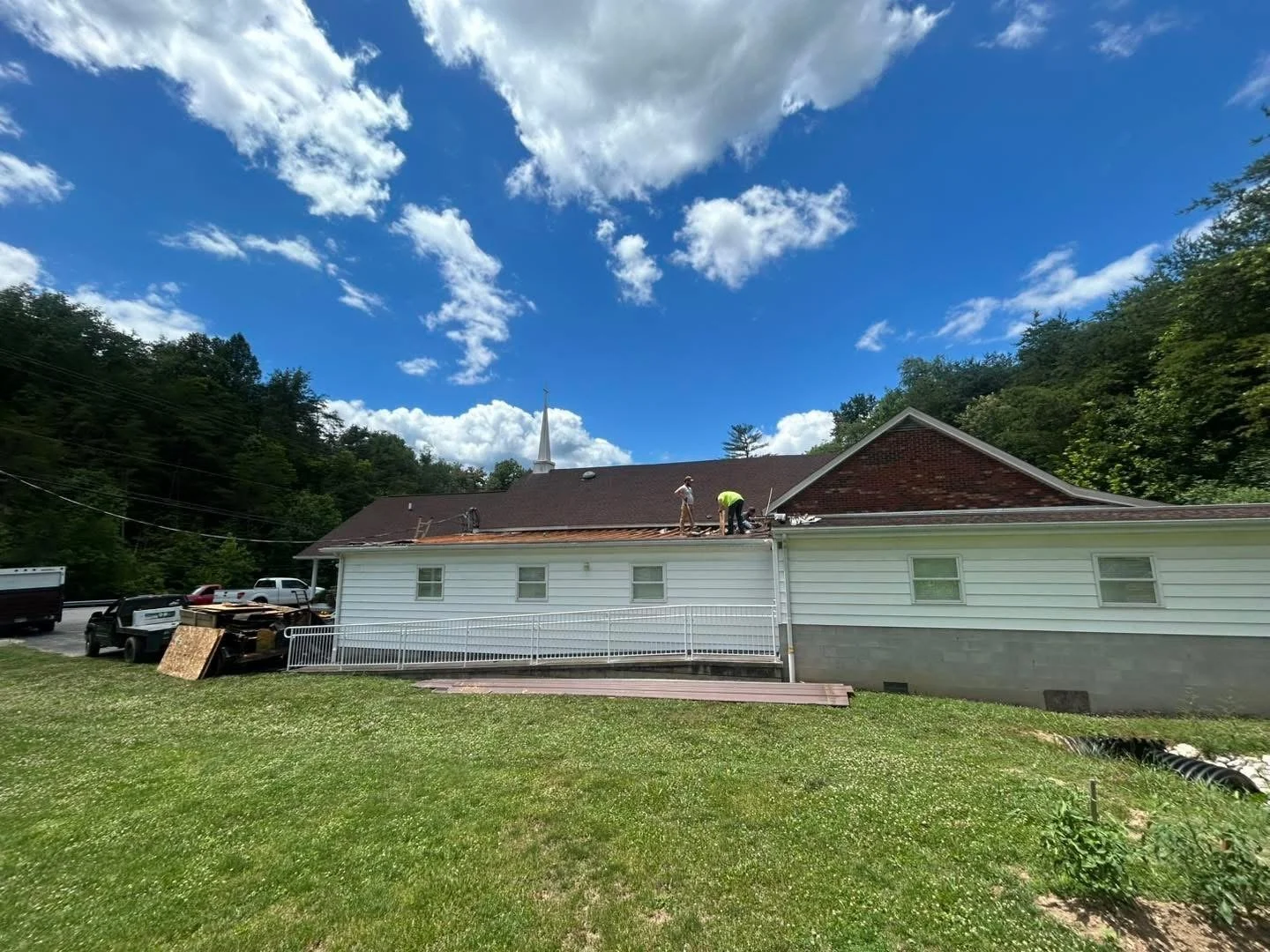 House undergoing roof repair under a blue sky with white clouds, workers on the roof, and parked vehicles nearby.