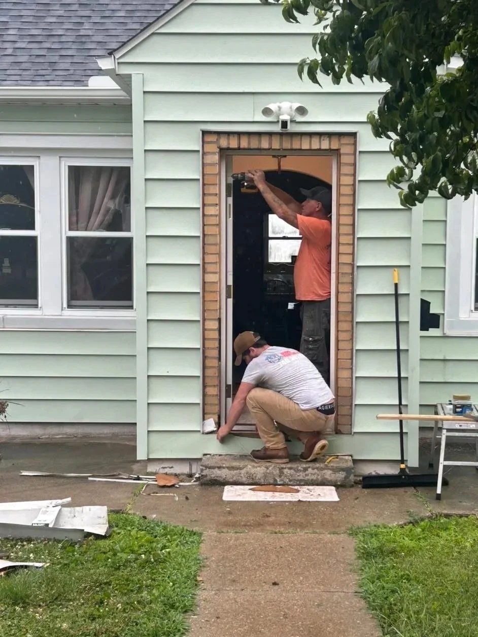 Two men installing a door frame on the front of a light green house, with one squatting and the other standing, working together. There are tools and materials nearby, including a long pole, a small table, and on the ground.