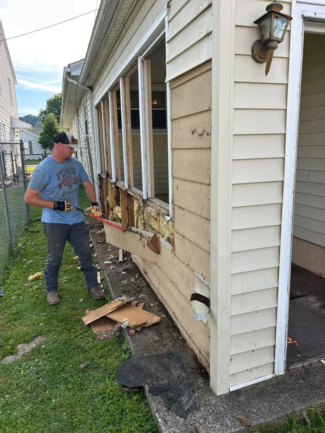 A man is tearing down the exterior wall of a house, exposing the inner wall and insulation. He is holding a crowbar, wearing gloves, jeans, and a t-shirt. Debris is scattered on the ground.