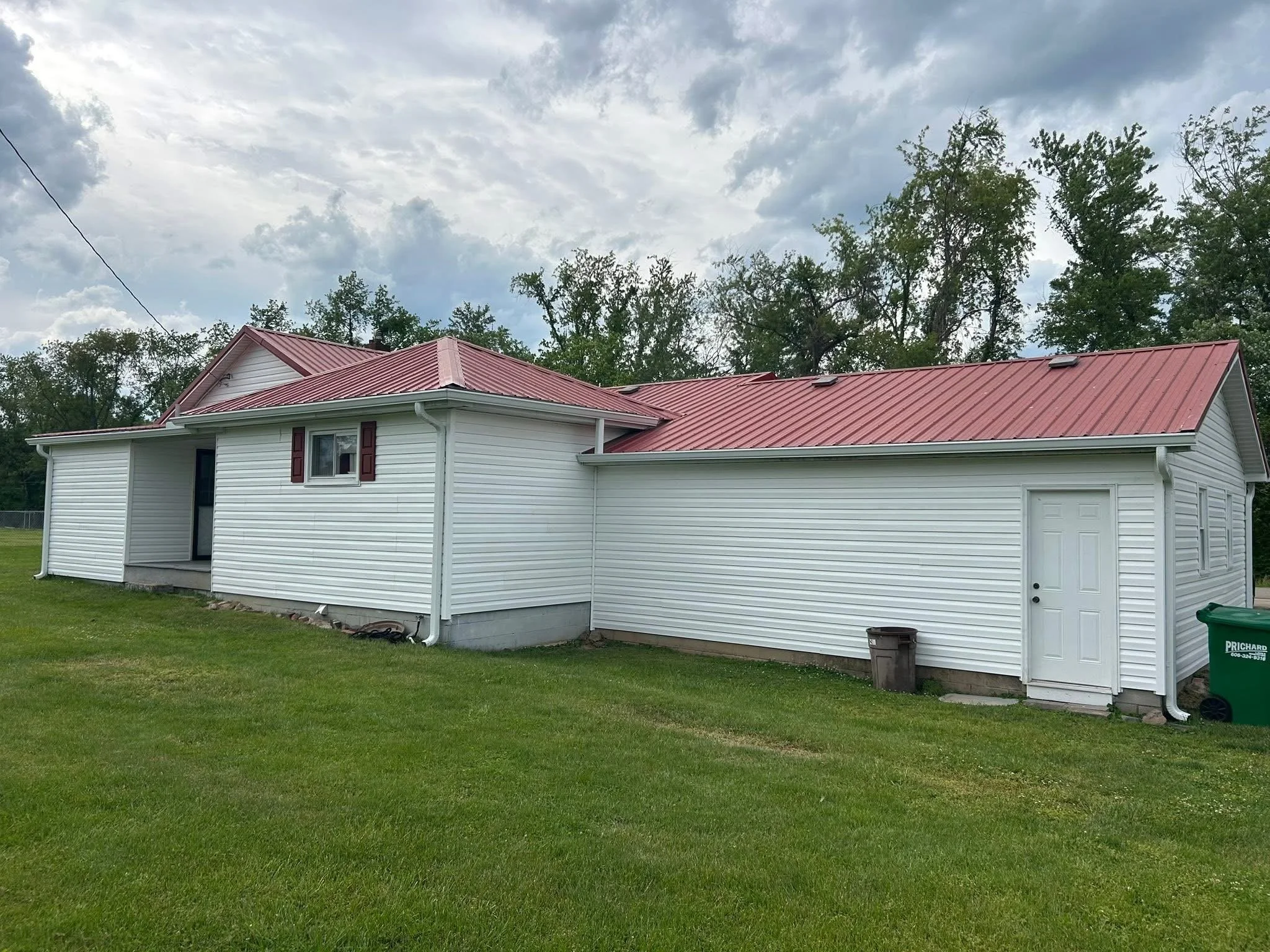 Back view of a house with white siding and a red metal roof, a garage door, and a small back door, with green grass and trees in the background, under a cloudy sky.