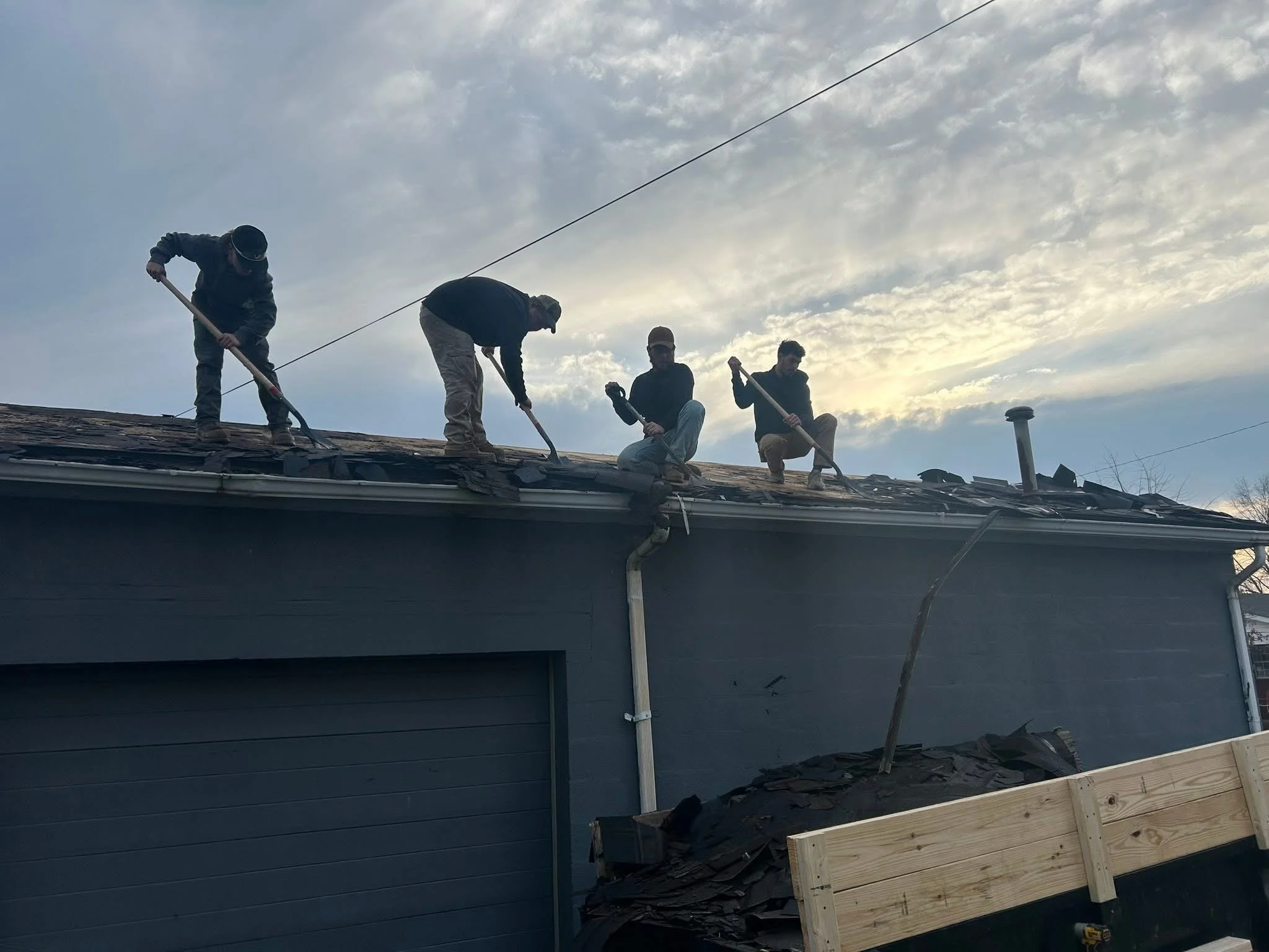 Four workers on a house roof replacing shingles during cloudy weather, with scaffolding and a wooden frame visible.