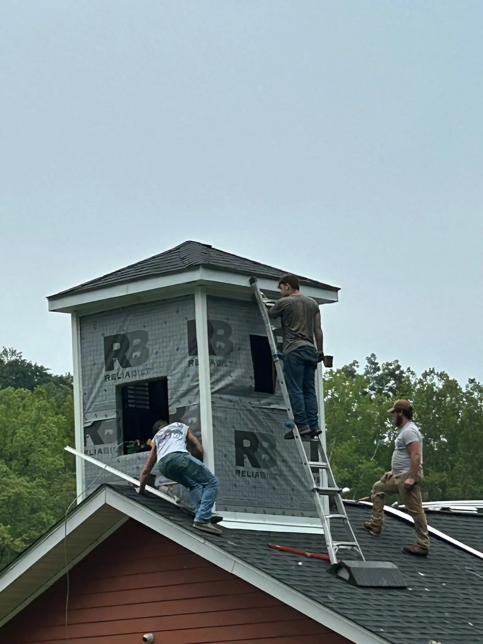 People working on the roof of a house during construction or repair, with some standing on the roof and others on a ladder, surrounded by trees and an overcast sky.