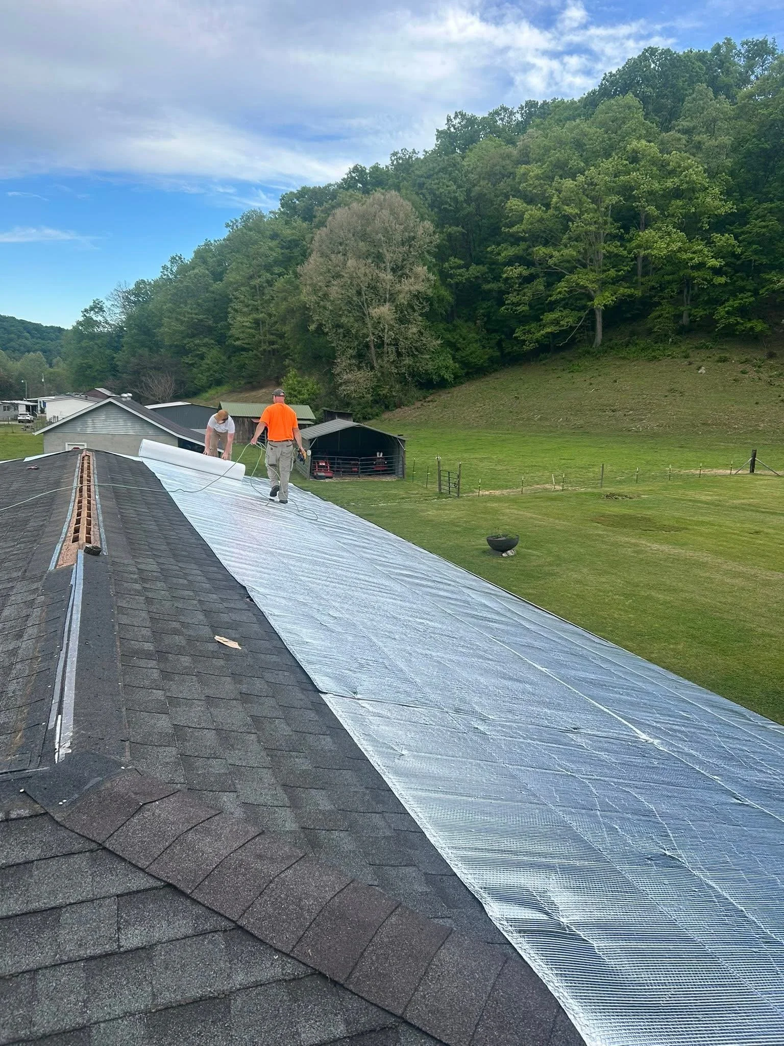 Two workers installing or repairing a metal roof on a residential building, with a landscape of trees and a grassy hillside in the background under a partly cloudy sky.