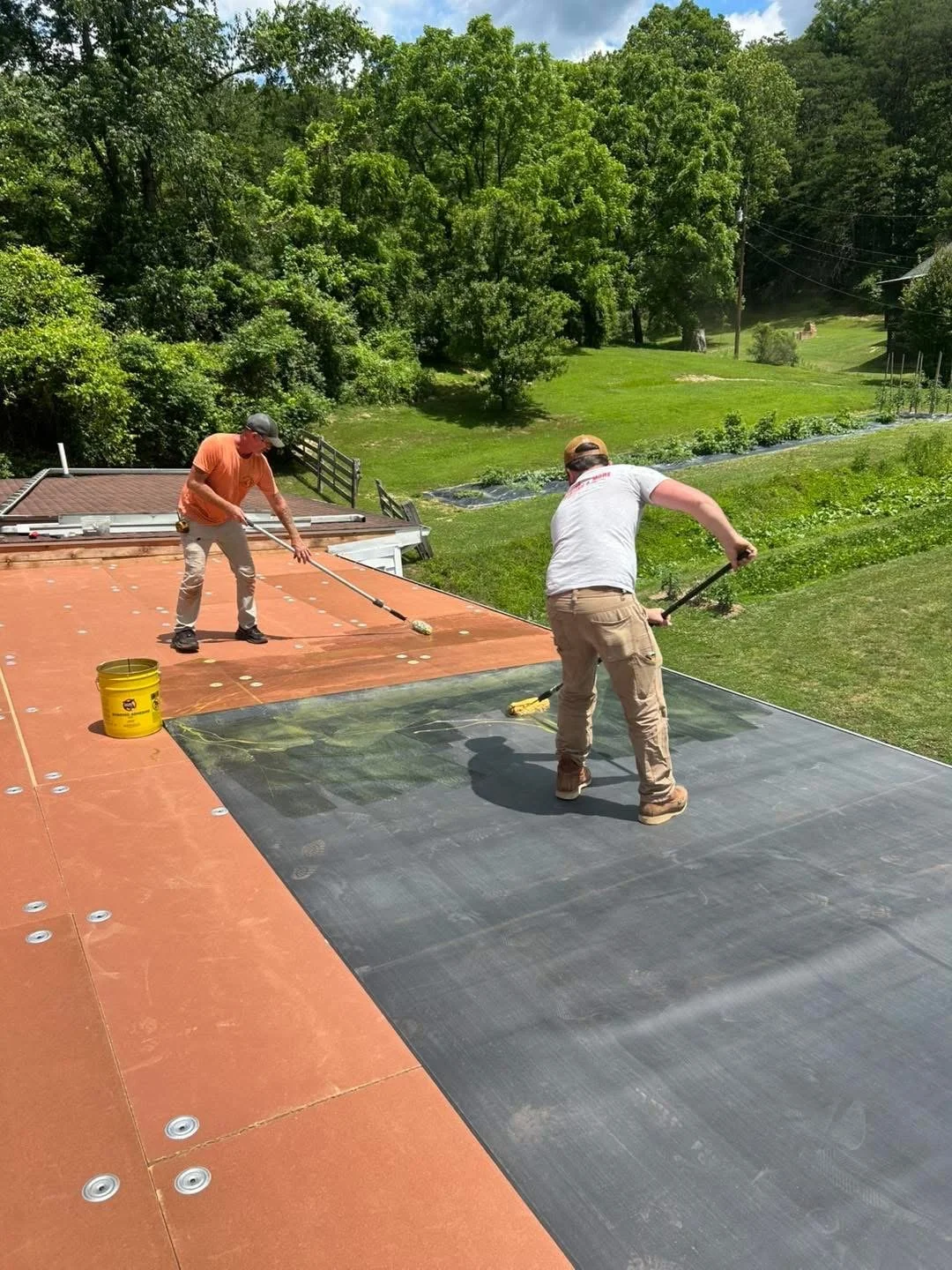 Two men working on installing a waterproof membrane on a flat roof, with green trees and grass in the background on a sunny day.