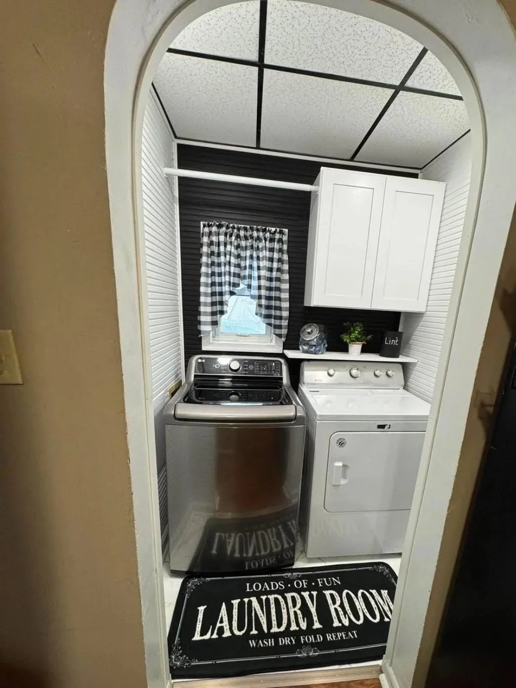 A laundry room with a washing machine and dryer, white cabinets, a checkered curtain, small window, and decorative rug.