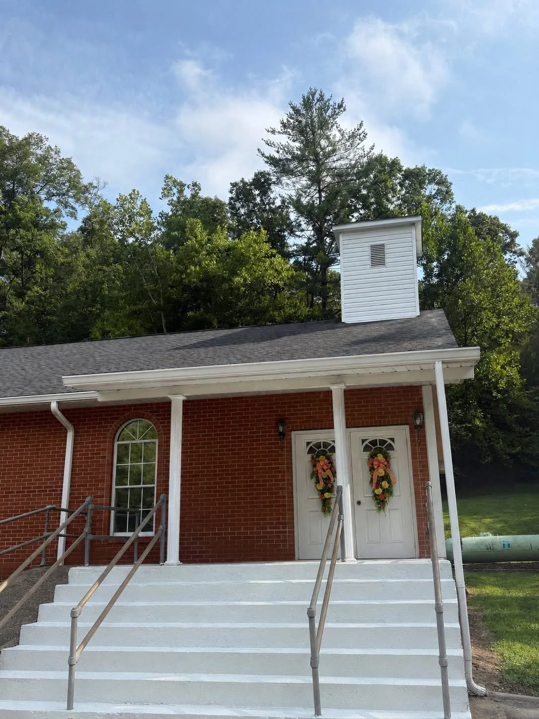 A brick church building decorated with two festive wreaths on white double doors, white stairs, and surrounded by green trees under a partly cloudy sky.