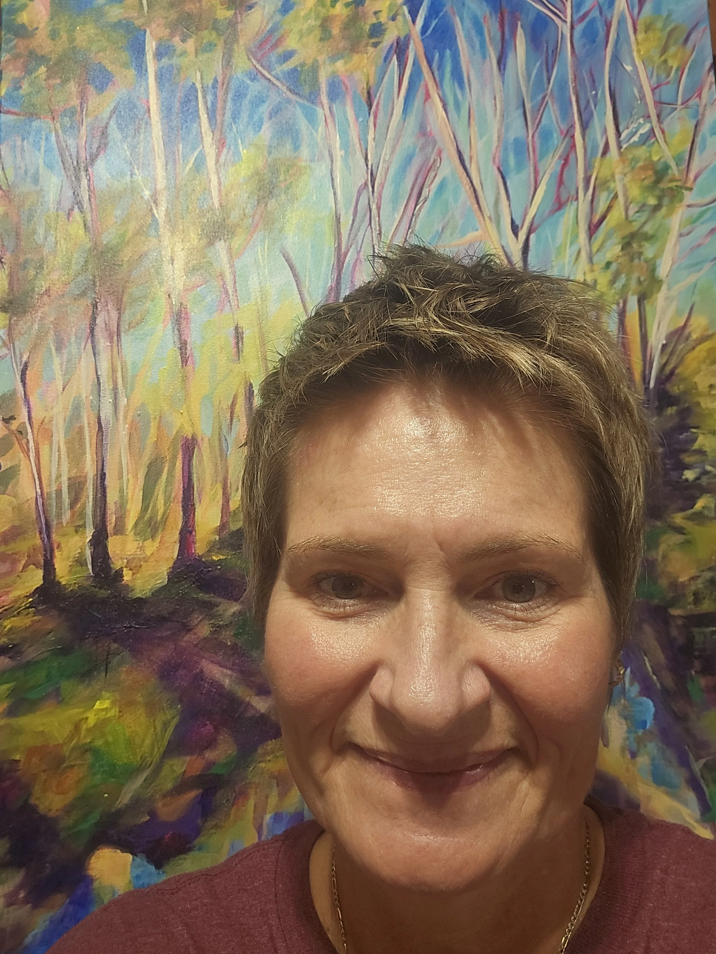 A woman with short, light brown hair and a gold chain necklace smiling in front of a colorful abstract painting of trees.
