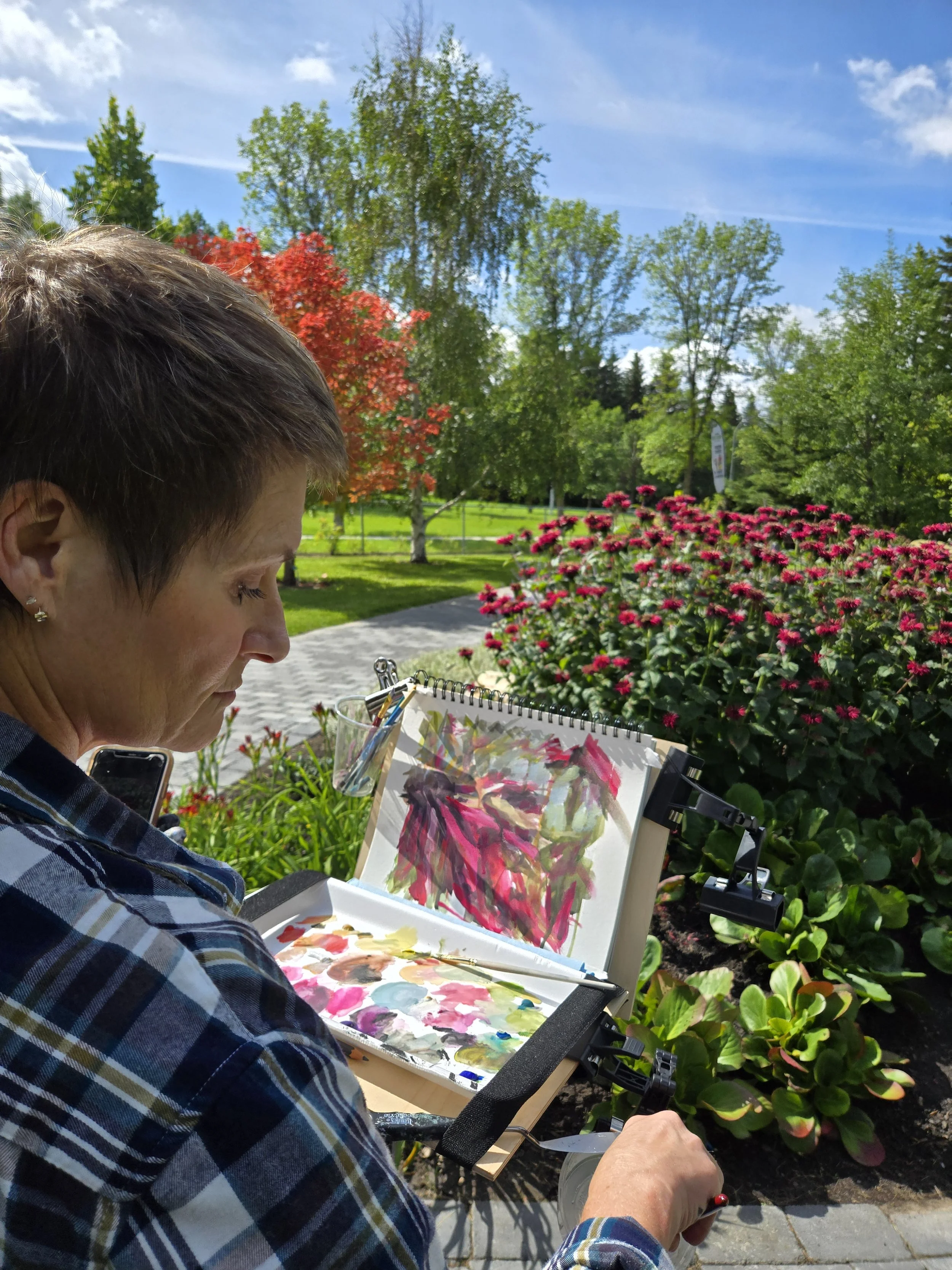 A woman painting outdoors on a bright, sunny day, with greenery and colorful flowers in the background.