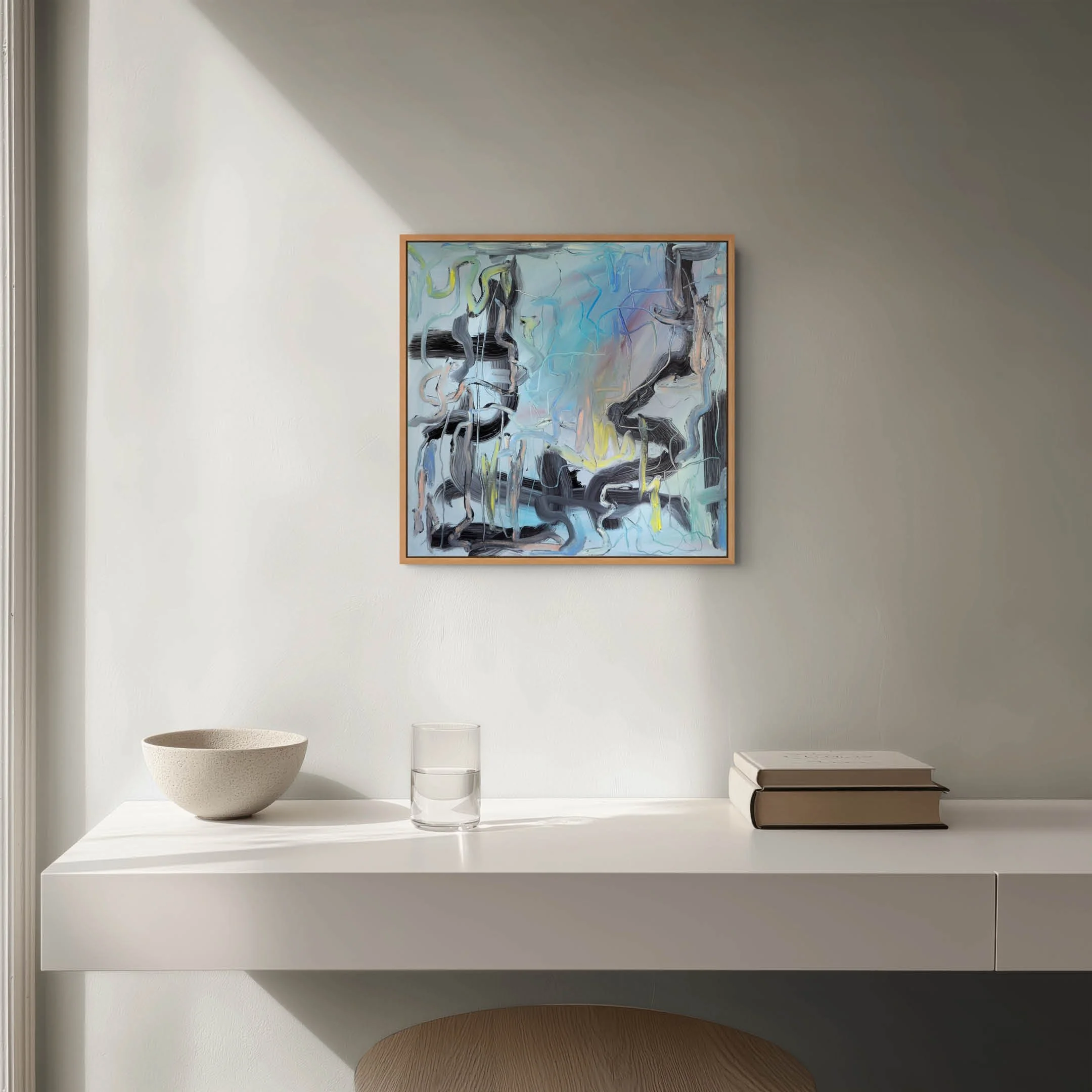 Minimalist white shelf with a white bowl, two clear glasses, and two books stacked horizontally. A framed abstract painting hangs on the wall above the shelf, and a wooden chair is partially visible at the bottom.