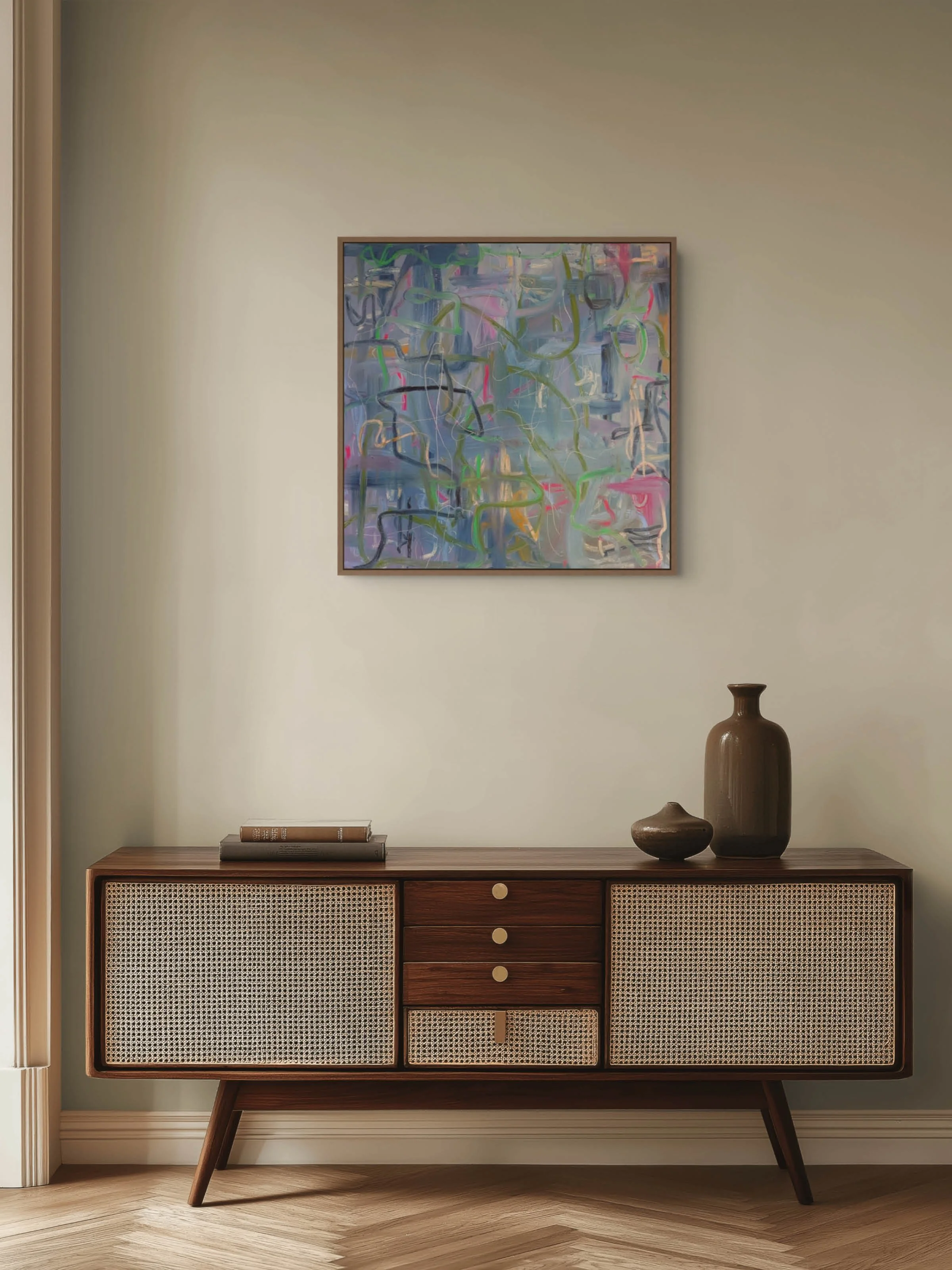A light-colored wall with a framed abstract painting hanging above a wooden sideboard. The sideboard features a woven rattan pattern on the cabinet doors and has three decorative vases and a stack of books placed on top.