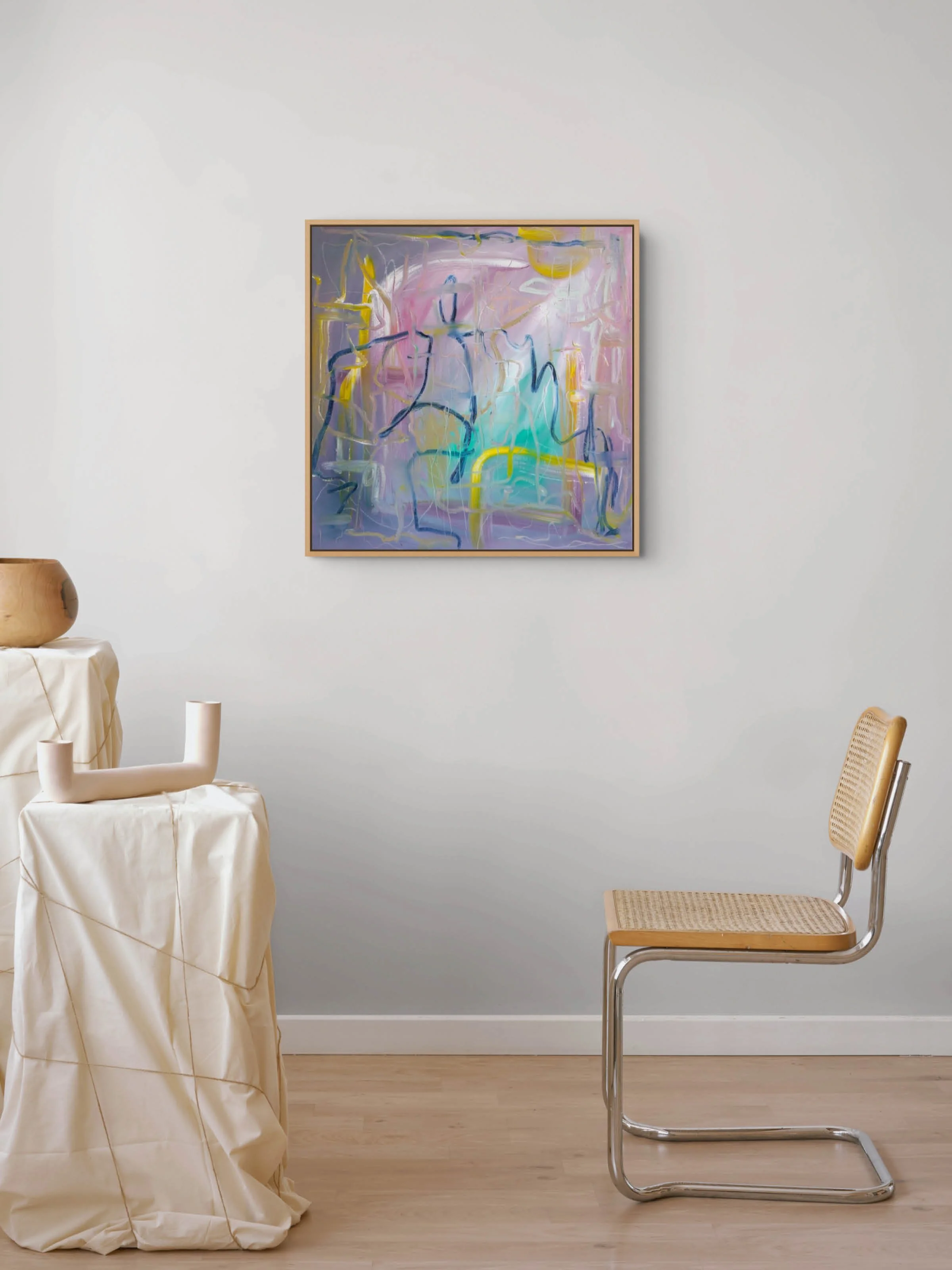 A room with a light gray wall, a modern wooden chair with a woven seat, a table covered with a cream-colored cloth, and a large abstract painting with pink, yellow, purple, and blue brushstrokes framed on the wall.