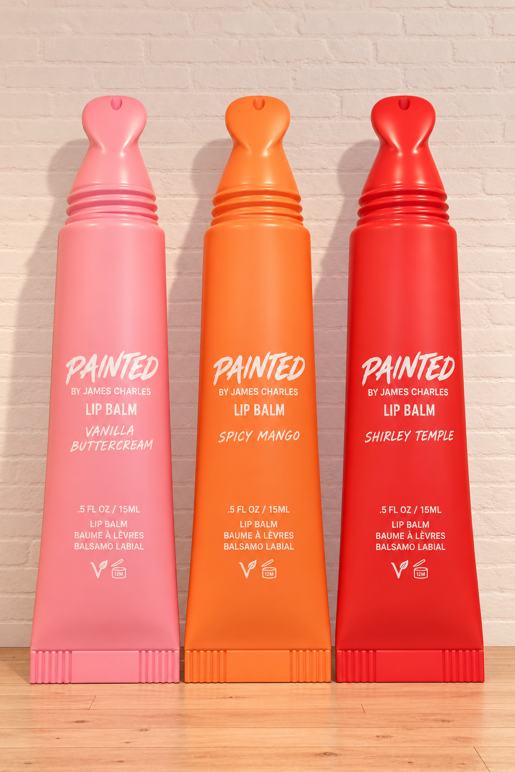 Three colorful tubes of lip balm in pink, orange, and red, labeled with different flavors, against a brick wall background.