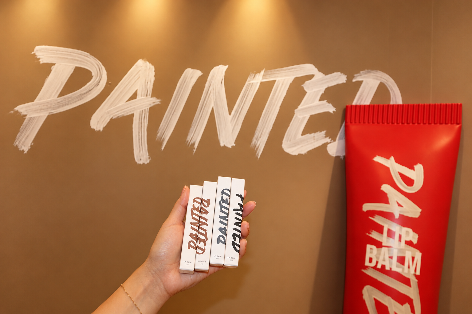 Hand holding four boxes of tinted lip balm in front of a wall with 'PAINTED' written in large white paint strokes, with a large red container or bag with white lettering on the right.
