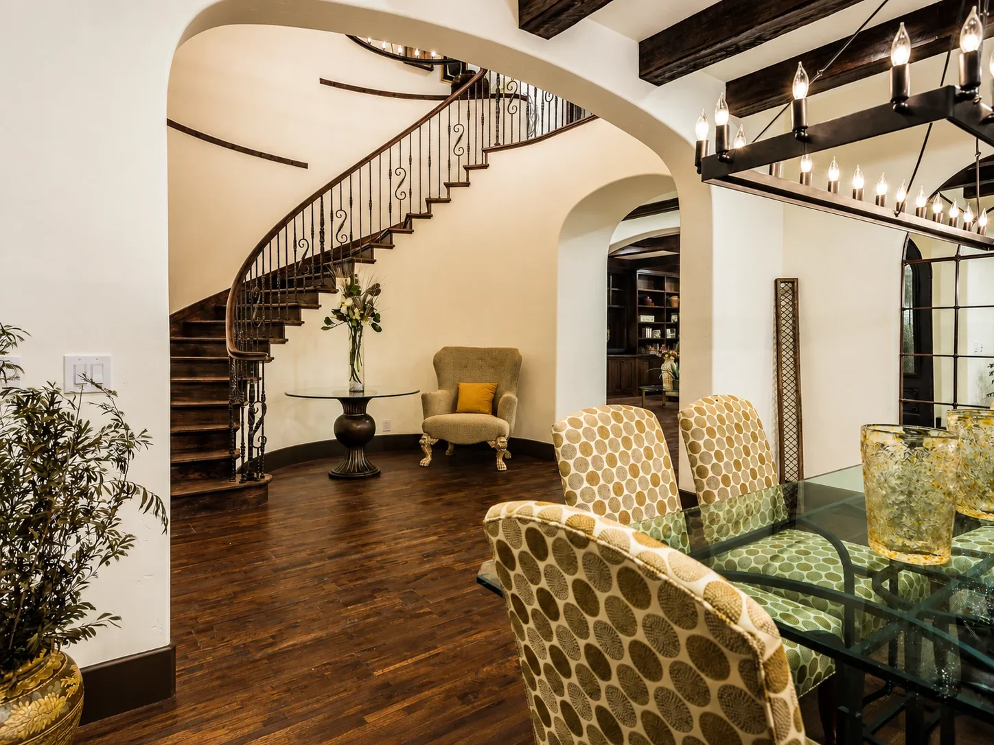 Dining and sitting area at Villa Englewood in Los Gatos featuring a wood staircase with iron railings, glass dining table, dark ceiling beams, warm wood flooring, and statement chandelier.
