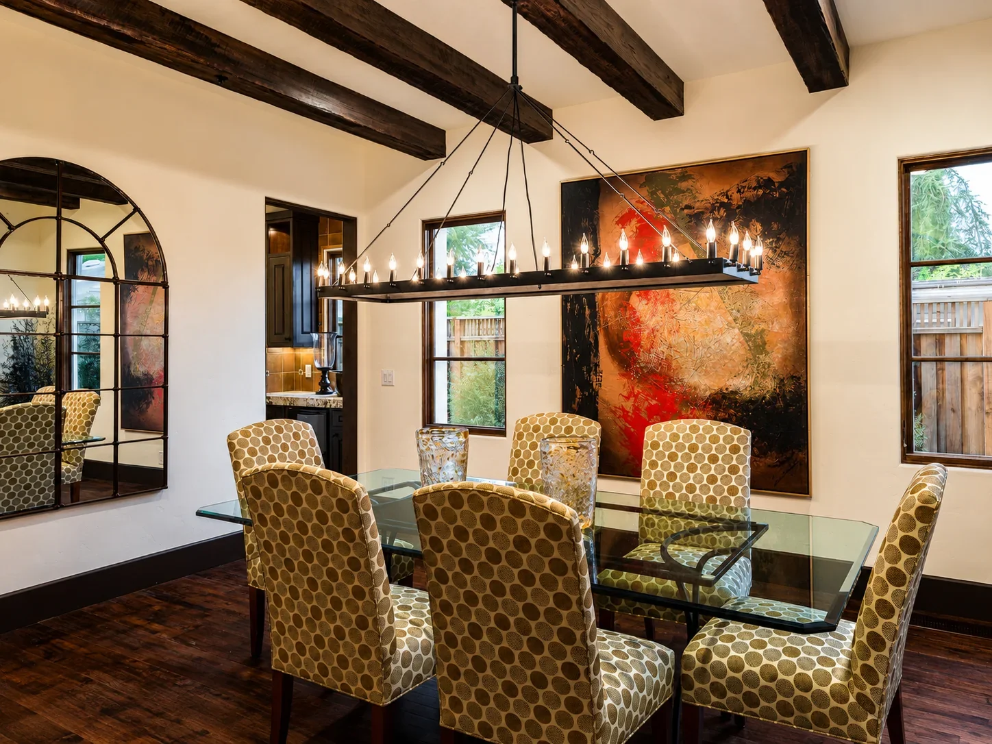 Dining room at Villa Englewood in Los Gatos featuring a glass dining table, patterned seating, abstract artwork, exposed wood beams, a candle-style chandelier, and garden-facing windows.