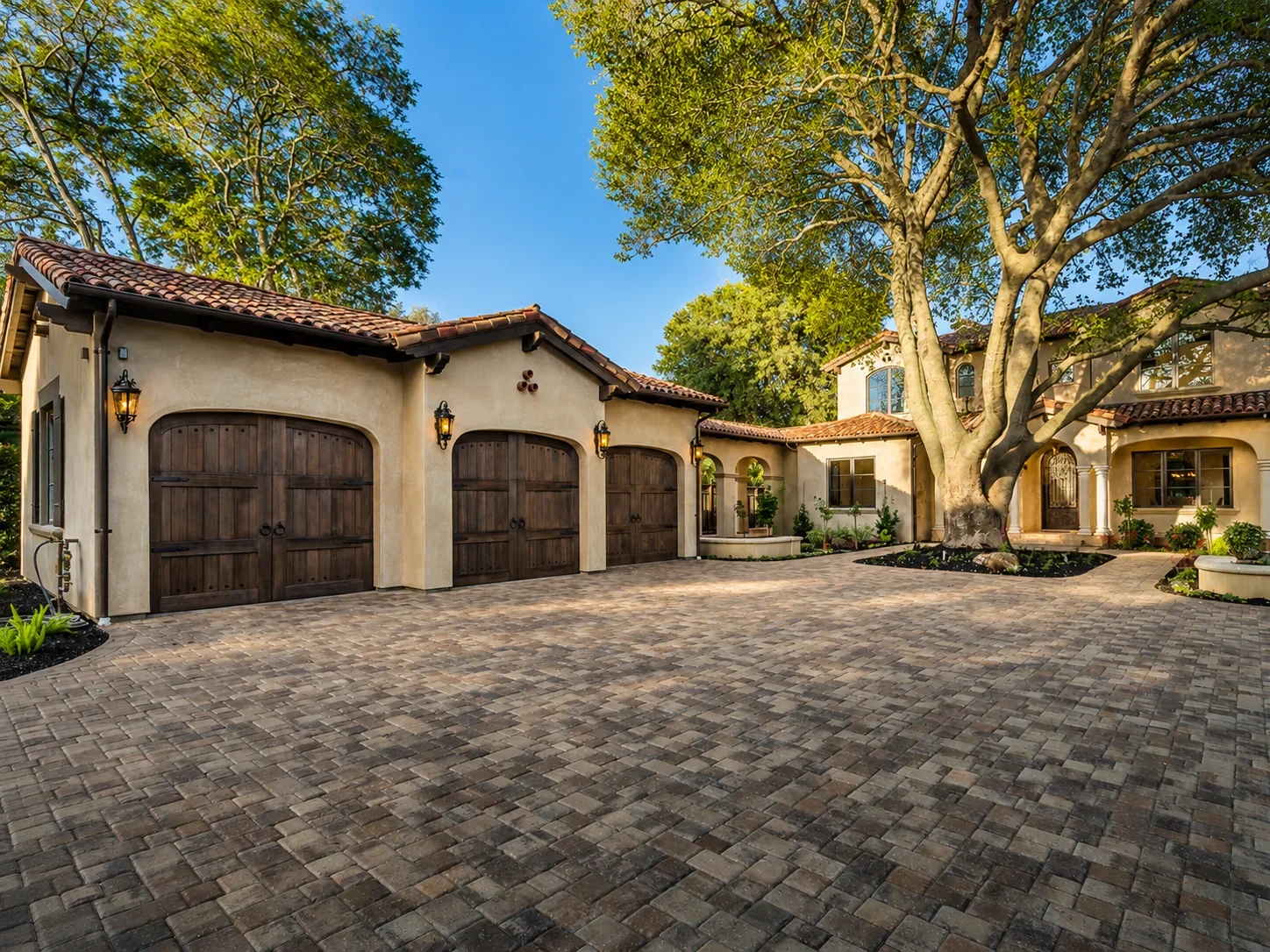 Front courtyard at Villa Englewood in Los Gatos, featuring the Spanish Revival residence, a stone-paved motor court, wood garage doors, lantern lighting, mature trees, and landscaped garden beds.
