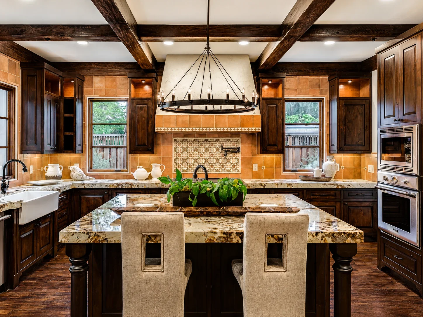 Kitchen at Villa Englewood in Los Gatos featuring dark wood cabinetry, a marble island with upholstered seating, a tile backsplash, and a central chandelier in a warm Spanish Revival setting.