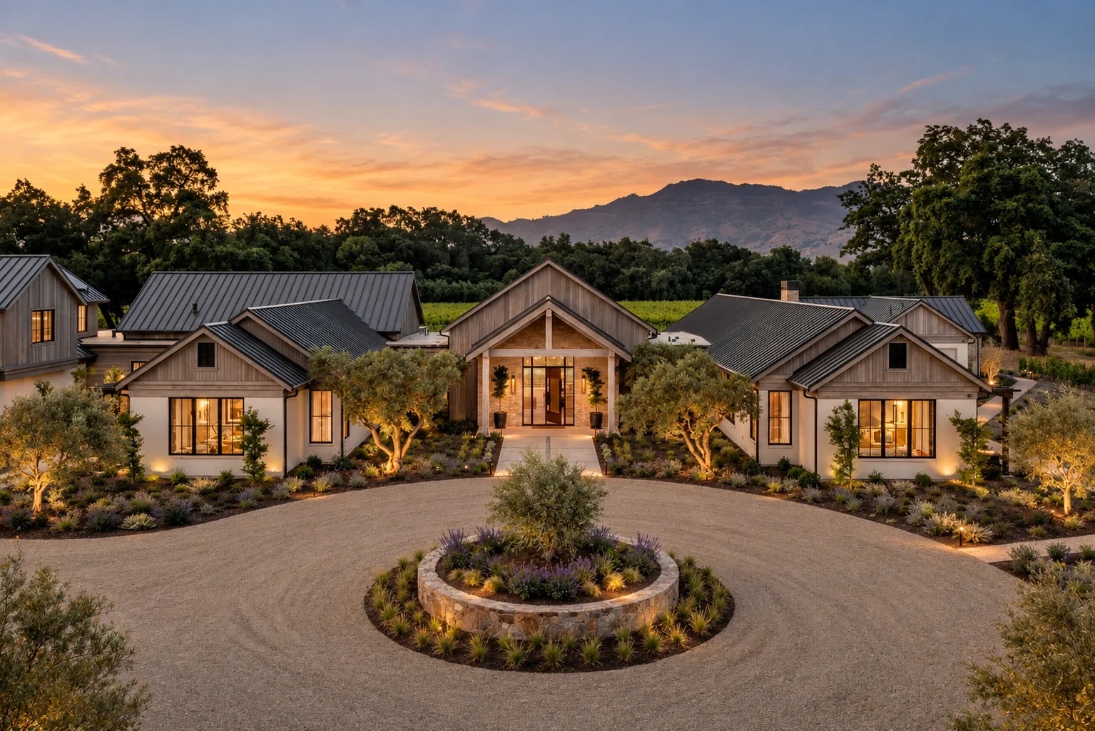 A modern house with a stone driveway, surrounded by landscaped gardens and trees, at sunset with mountains in the background.