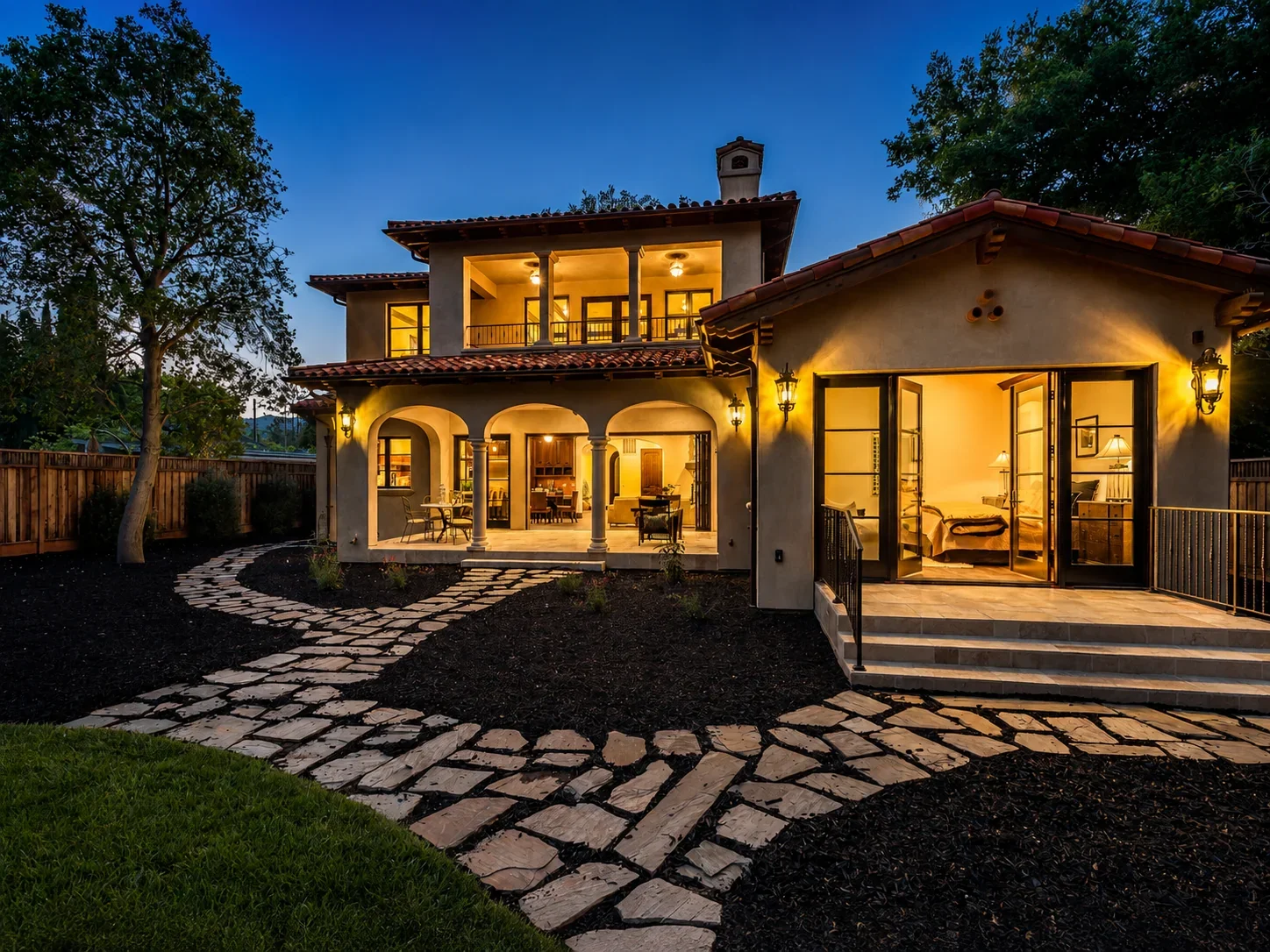Dusk exterior of Villa Englewood in Los Gatos, featuring the two-story Spanish Revival residence, arched windows and doors, a stone pathway, mature landscaping, and warm interior light.