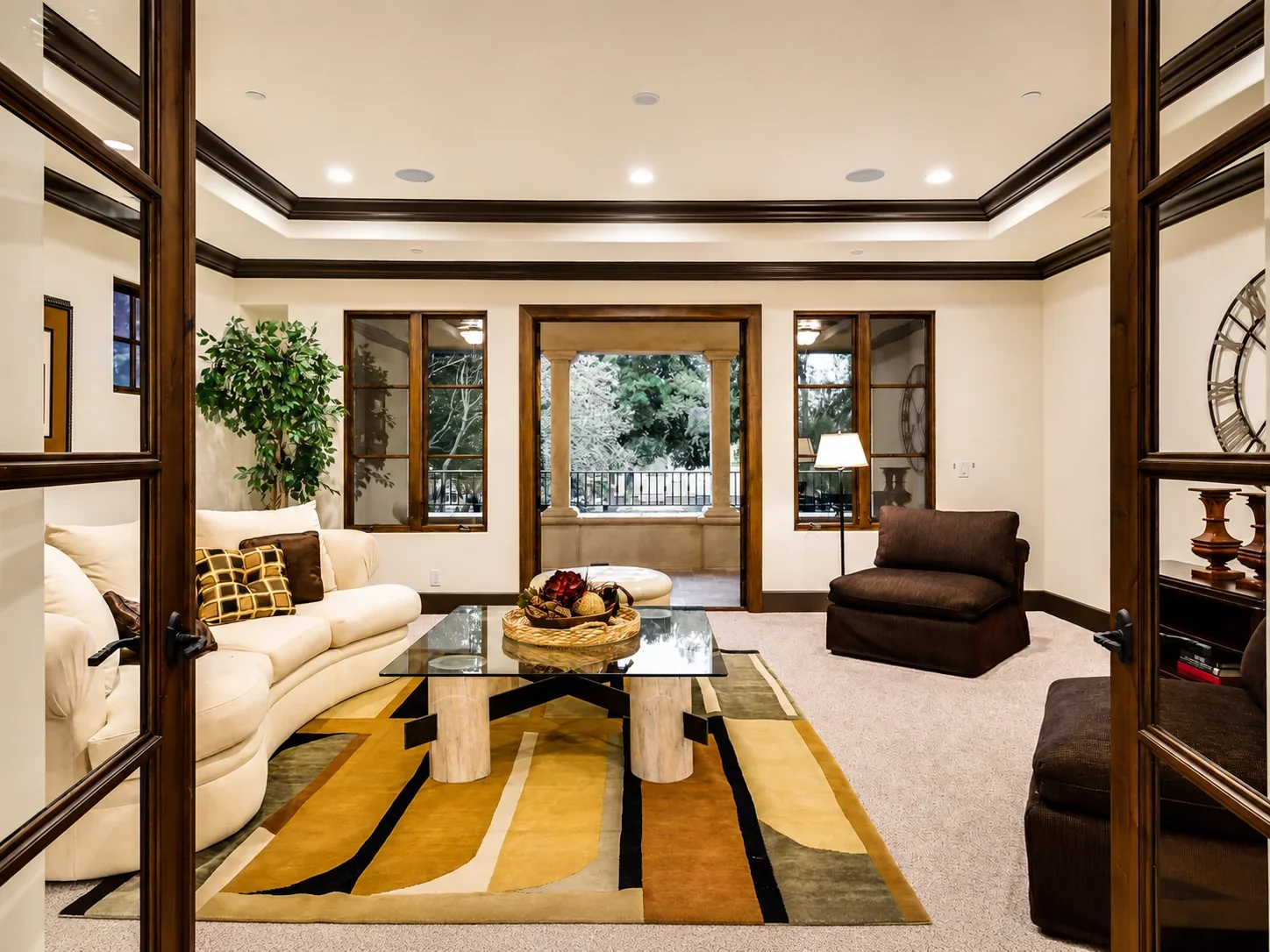 Living room at Villa Englewood in Los Gatos featuring a cream sofa, brown lounge chairs, a glass coffee table, patterned rug, wood trim, and expansive windows overlooking the landscaped exterior.
