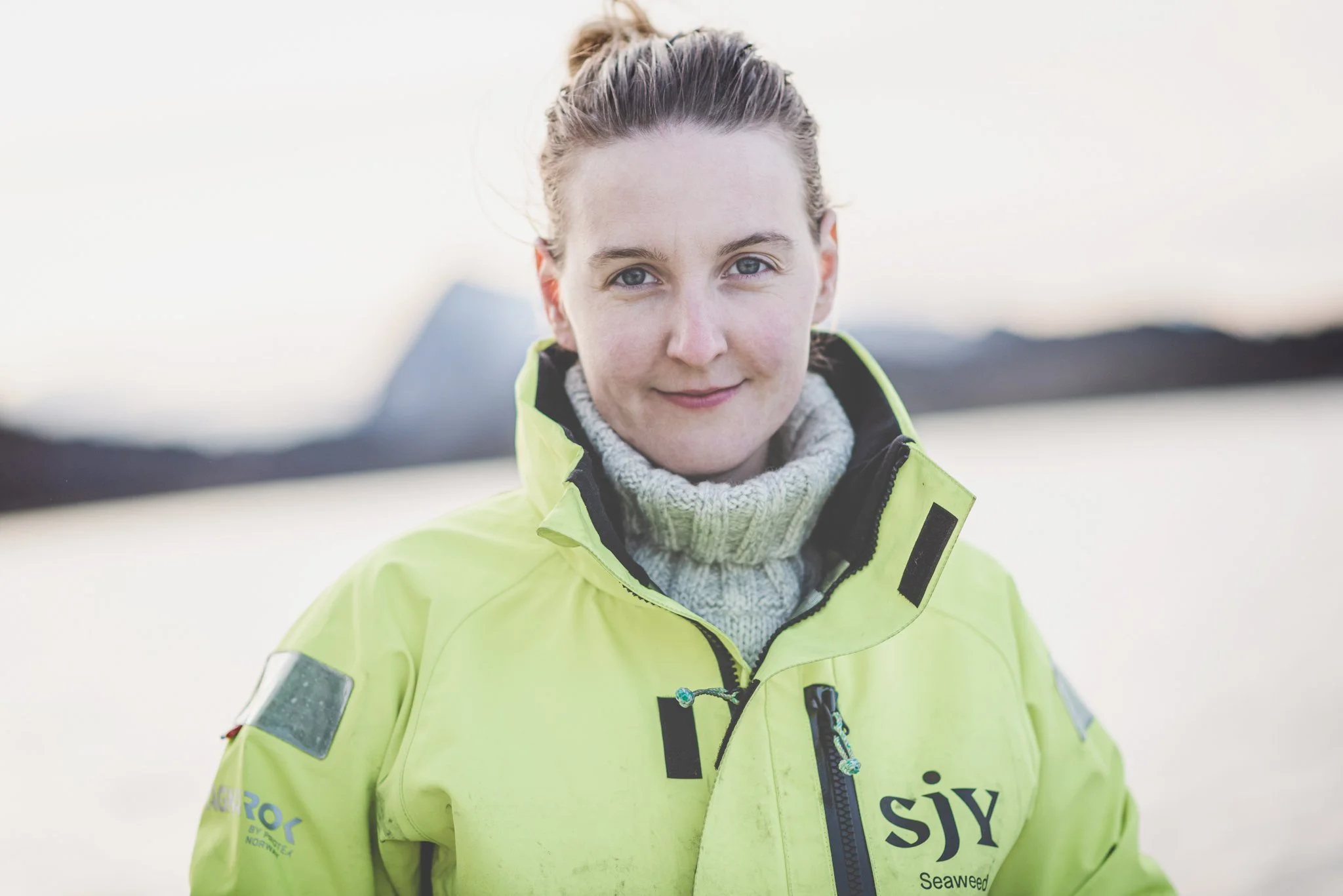 A woman wearing a bright yellow jacket and a gray turtleneck sweater outdoors, smiling with blue eyes and light hair tied back, with a blurred background of snow and terrain.