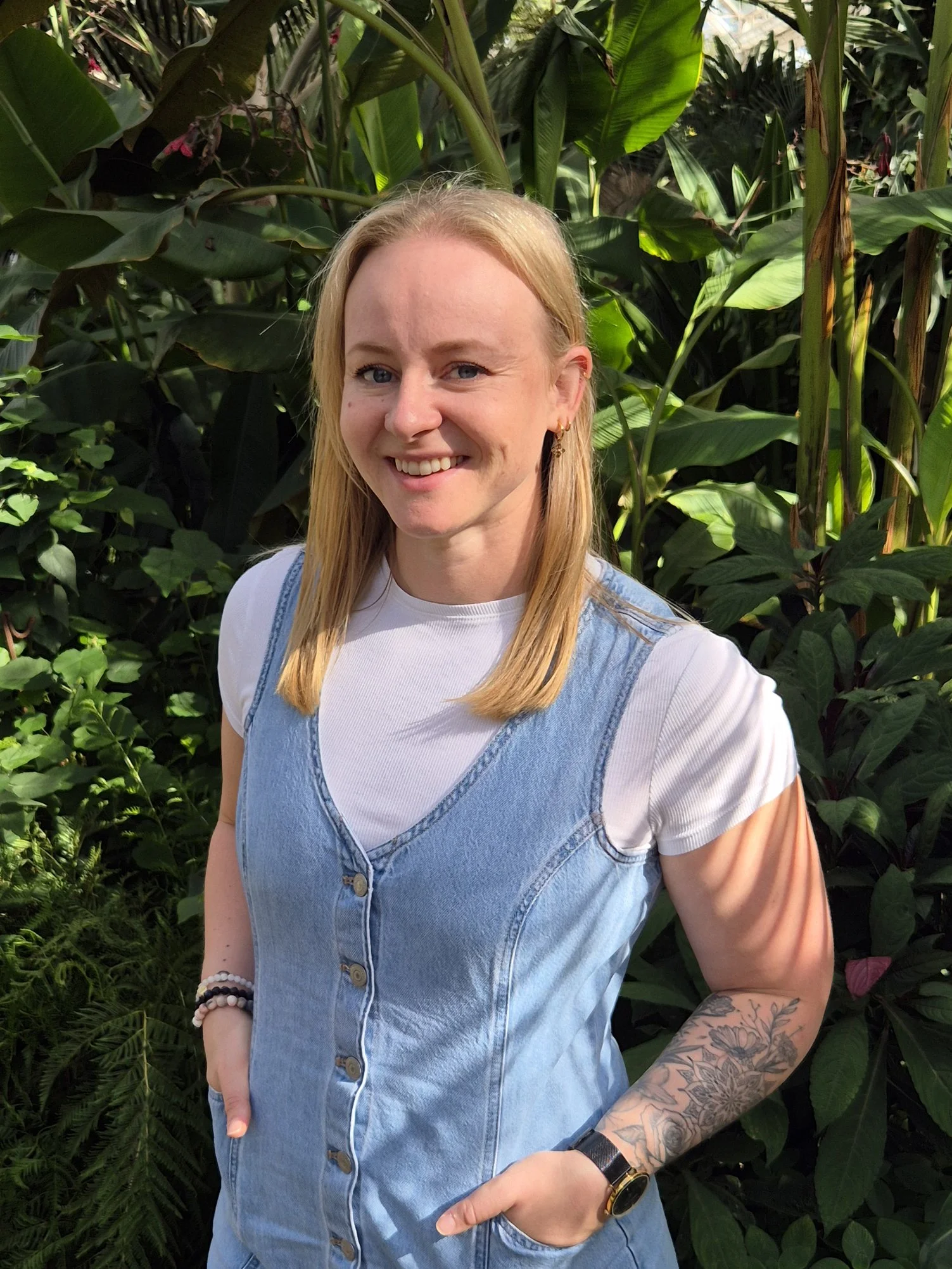 A woman with blonde hair wearing a white shirt and denim vest, standing among green foliage, smiling at the camera, with tattoos and bracelets visible on her arm.